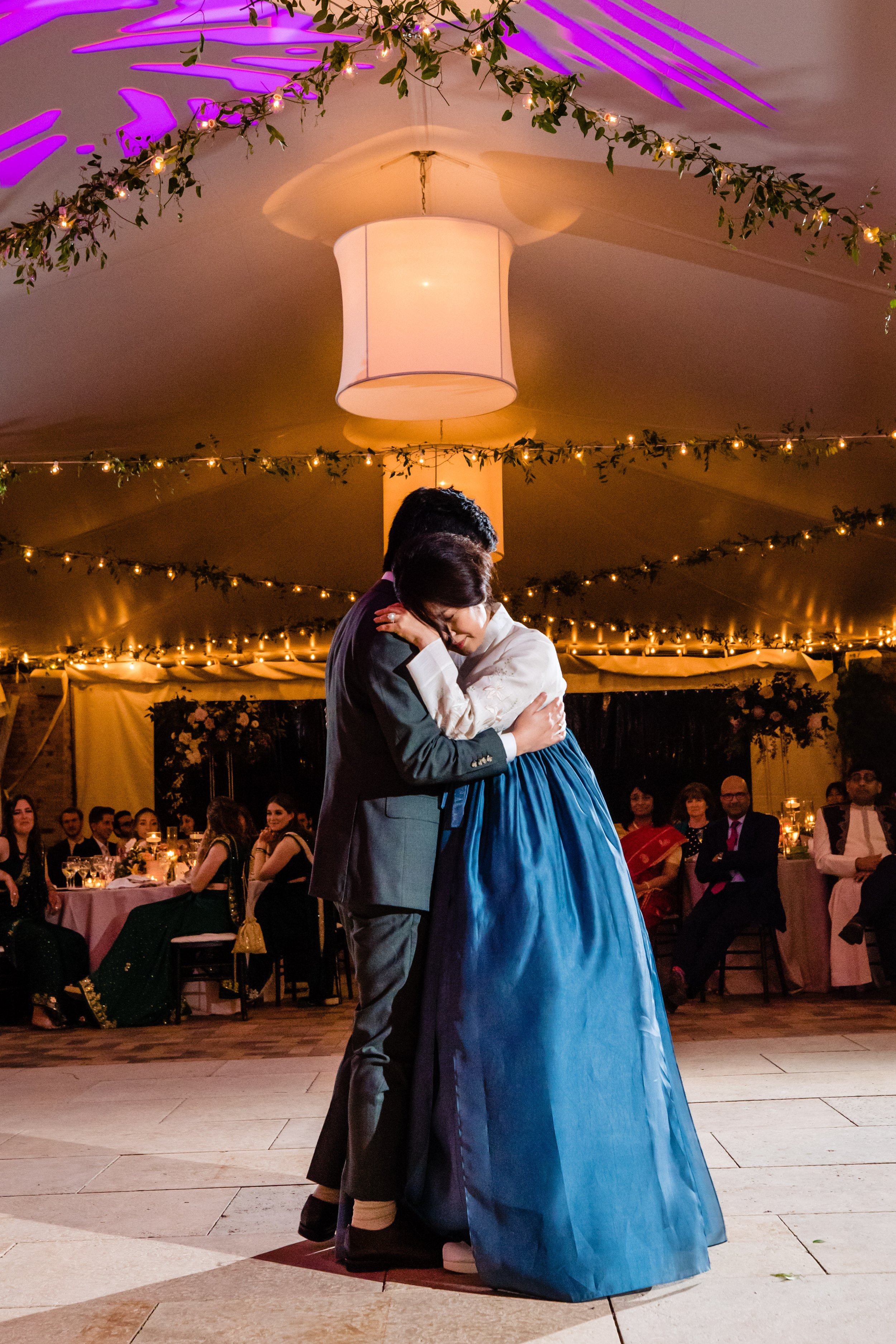 A couple is sharing a kiss on a dance floor during a wedding reception, with guests watching and seated at decorated tables in the background.