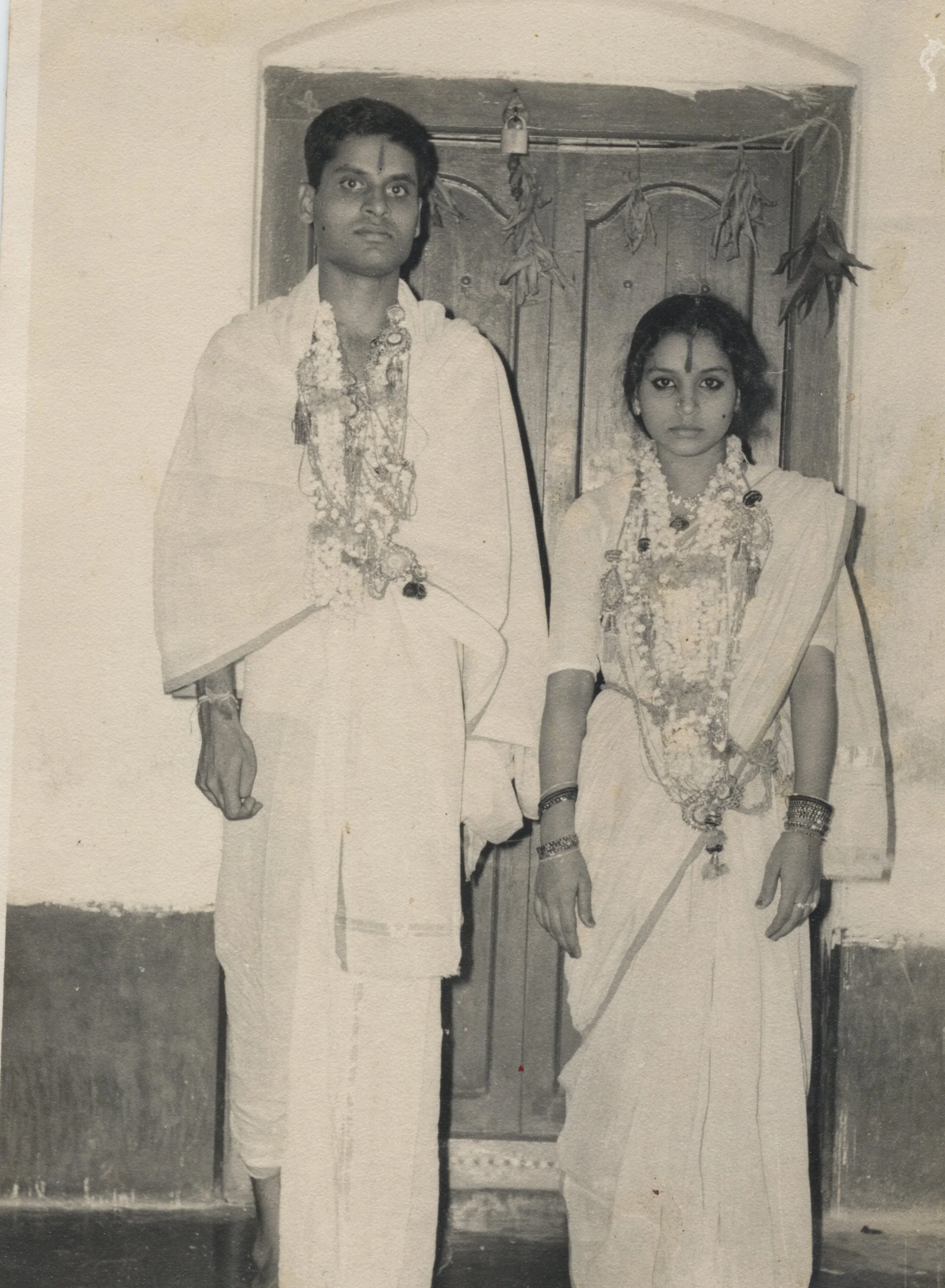 A black-and-white photo of a man and woman in traditional Indian attire, standing in front of a door with hanging dried leaves or herbs. The man is taller, dressed in a white kurta with multiple necklaces, and the woman is wearing a saree with jewelry and has her hands at her sides.