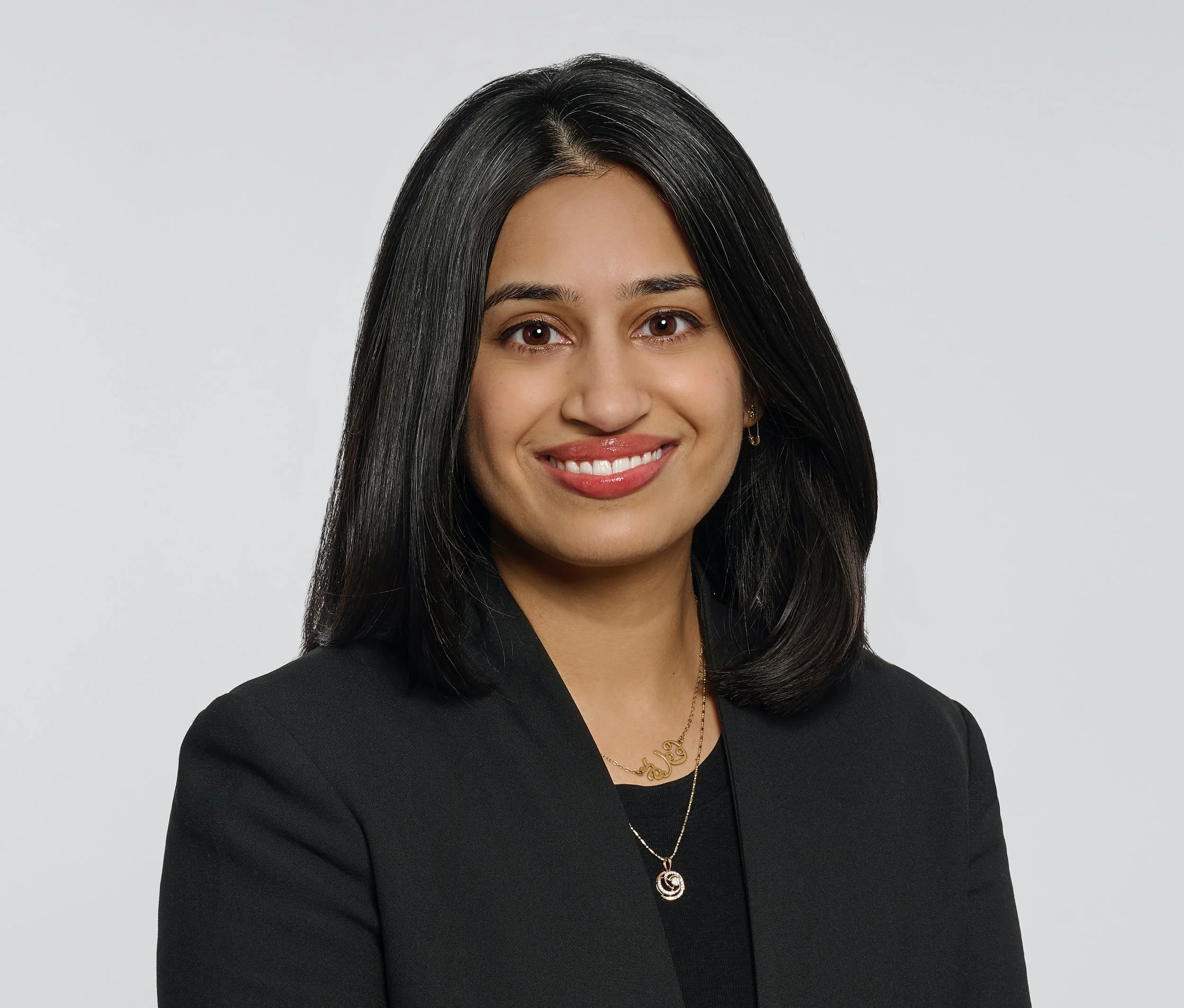 Portrait of a woman with long, straight black hair, wearing a black blazer, gold jewelry, and smiling at the camera against a plain light background.