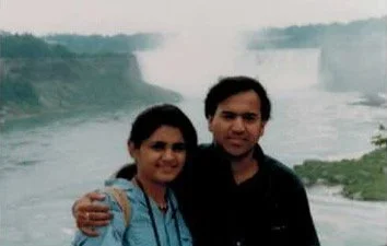 A man and woman standing next to each other outdoors near a large waterfall, smiling at the camera.
