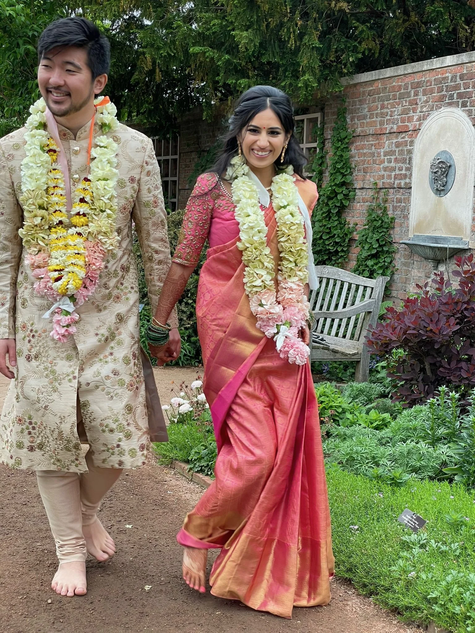 A man and a woman walking outdoors in a garden, holding hands, dressed in traditional Indian attire, wearing flower garlands, smiling, with greenery and a brick wall in the background.