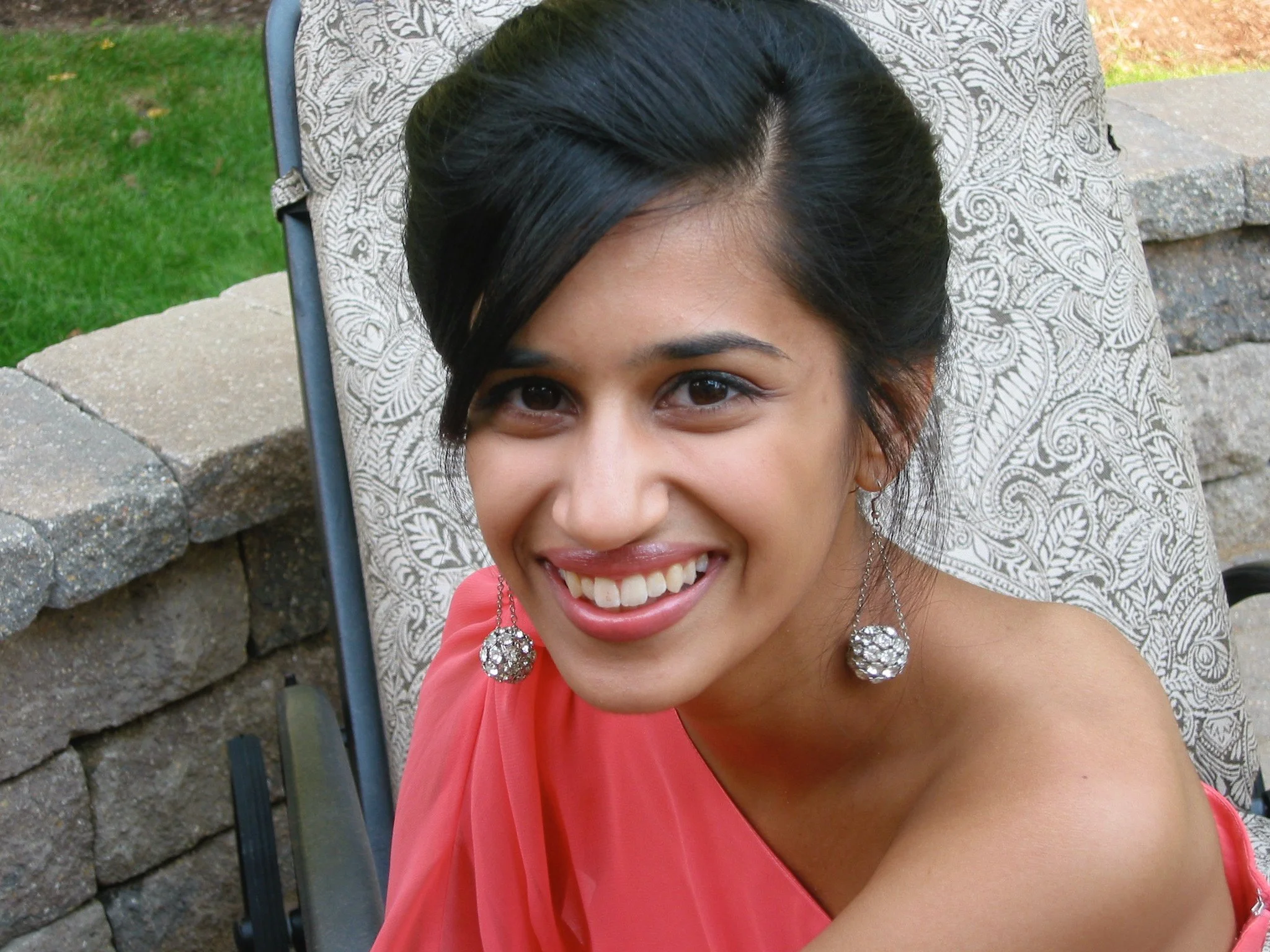 Close-up of a young woman smiling, sitting outdoors on a chair with a decorative patterned fabric. She has dark hair styled back, wearing large earrings with crystal embellishments, and a coral dress.