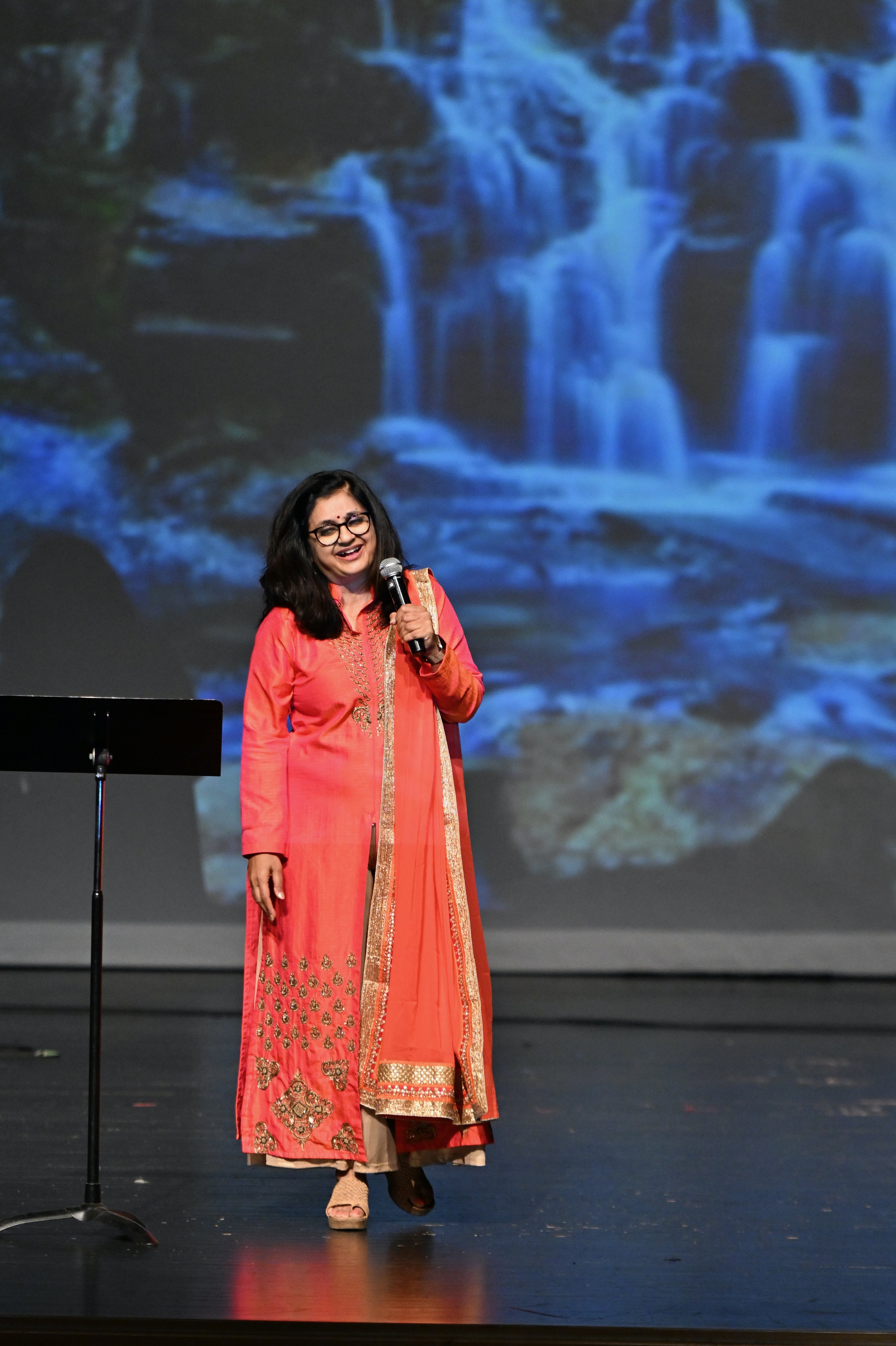 A woman with black hair and glasses wearing a traditional orange and gold Indian dress, standing on stage holding a microphone, with a backdrop of a blue waterfall scene.