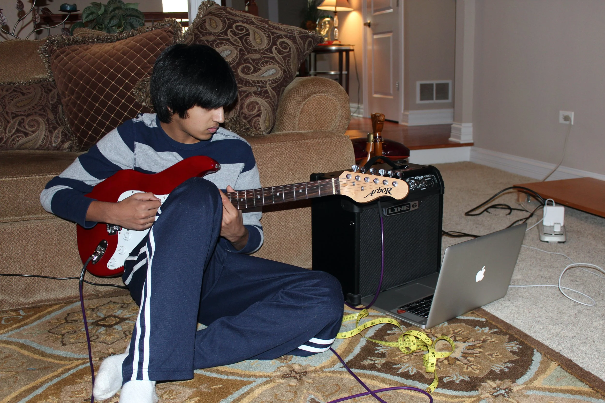 A young person with black hair sitting on the floor playing an electric guitar, wearing a striped long sleeve shirt and sweatpants, surrounded by electronic equipment including a laptop, guitar amplifier, and measuring tape. The setting appears to be a living room with furniture, a rug, and a lamp in the background.