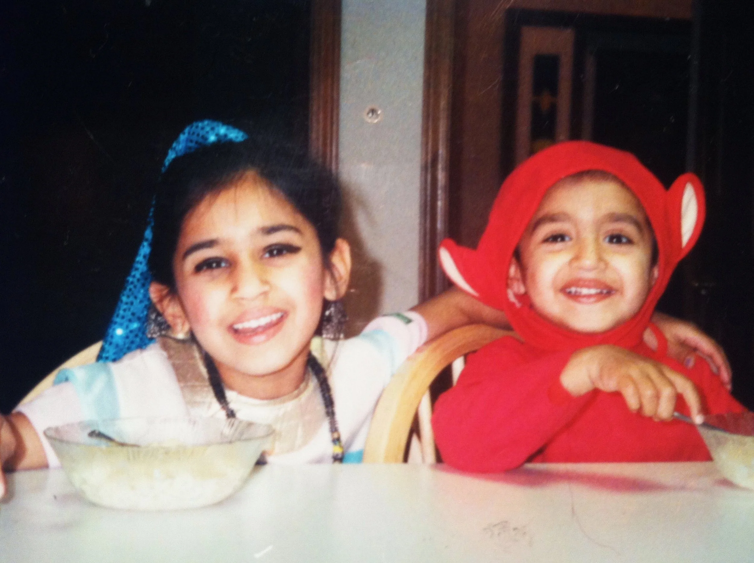 Two children, a girl and a boy, smiling and dressed in costumes. The girl is wearing a white shirt with colorful decorations and a blue headband, while the boy is dressed as a toy soldier with a red hat and outfit.