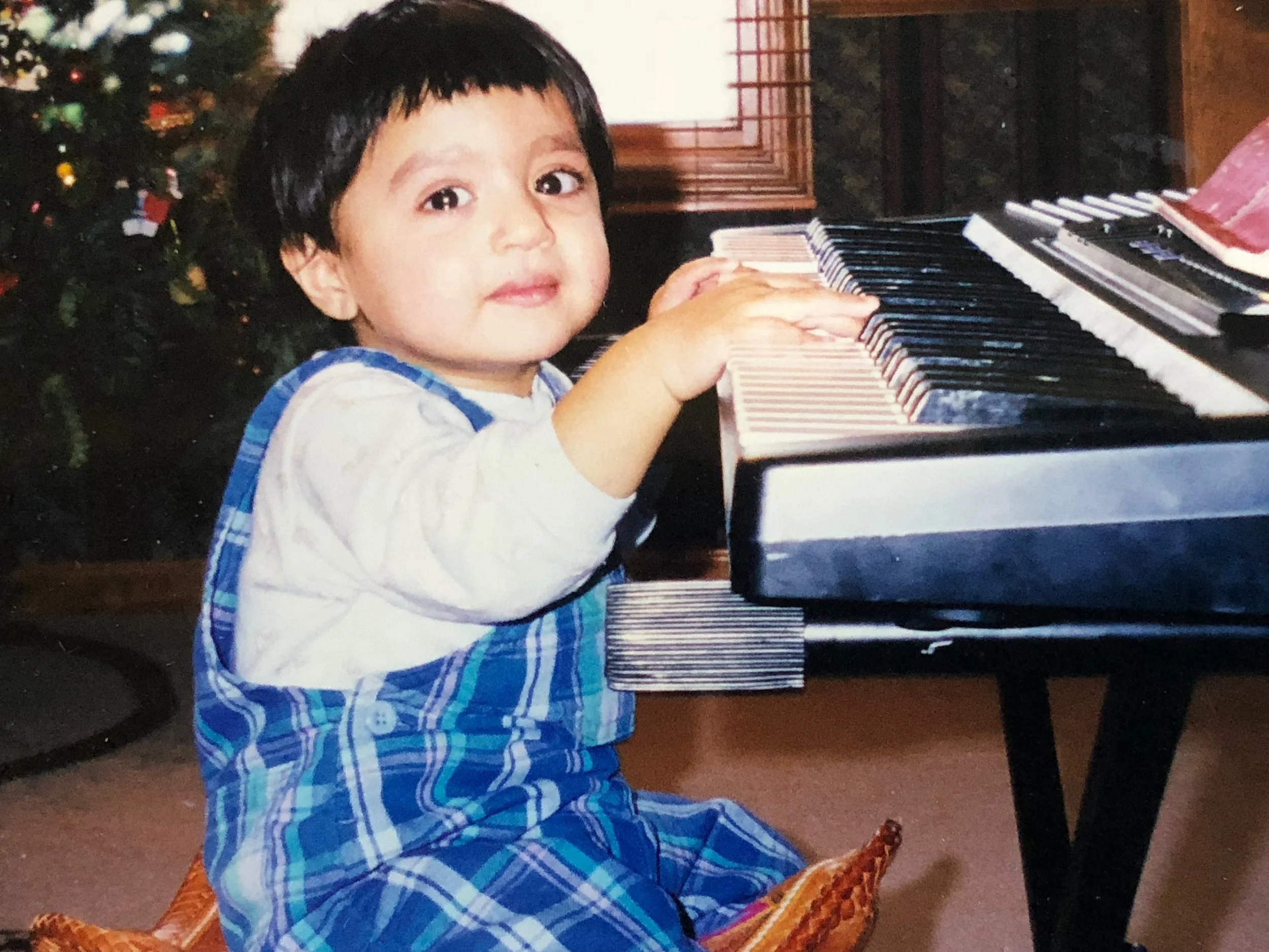 A young child with short dark hair and light skin, wearing a blue plaid shirt, sitting on a chair and playing a piano. A Christmas tree with ornaments is in the background.