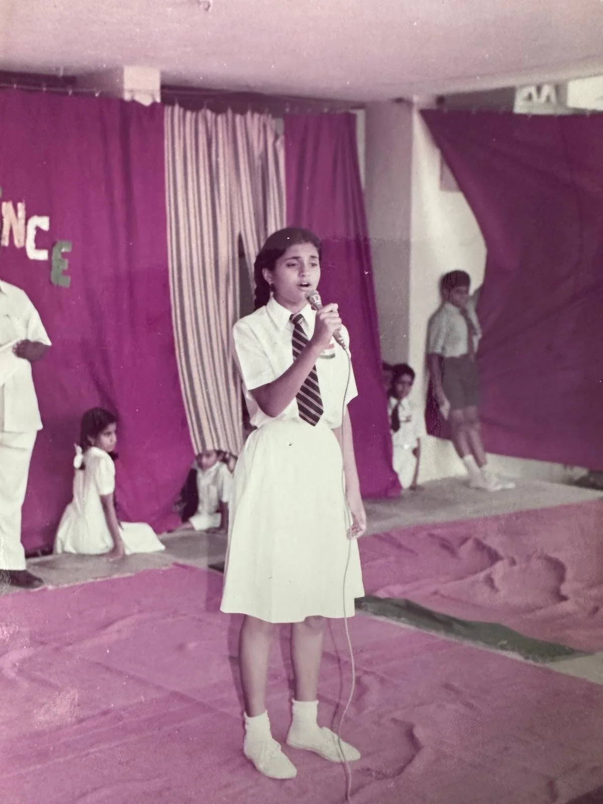 A young girl in a school uniform holding a microphone on stage, with other children in school uniforms sitting and standing in the background, during a school performance or event.