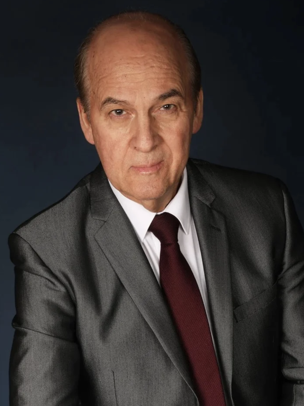 A middle-aged man in a dark grey suit, white shirt, and maroon tie, posing against a dark background.