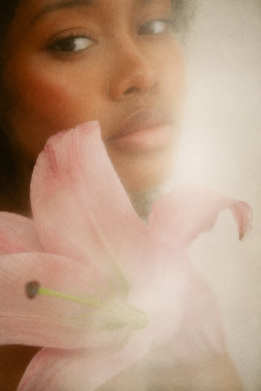 A woman with dark skin and curly black hair holding a pink lily flower close to her face.