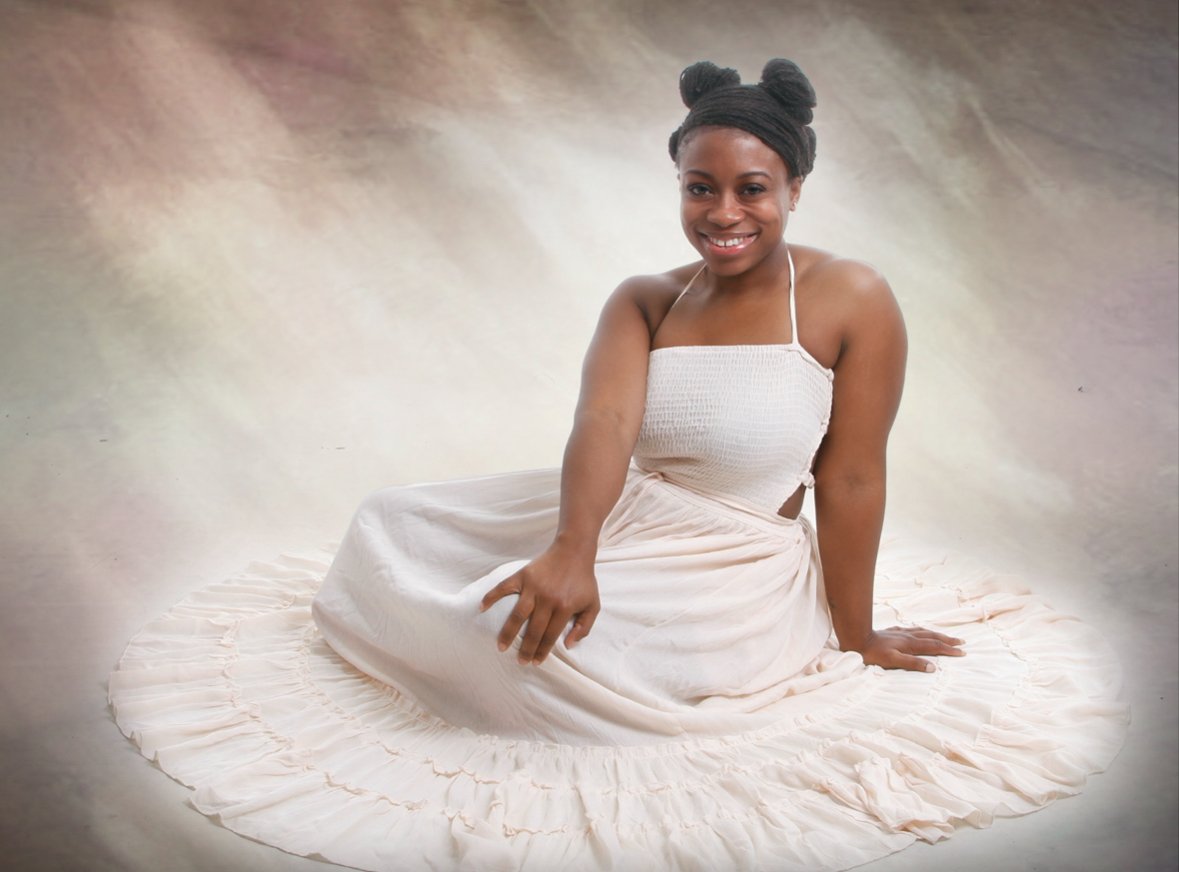 A young woman with dark skin and black hair styled in buns, wearing a white strapless dress, sitting on the floor with a gentle smile against a soft, blurred background.