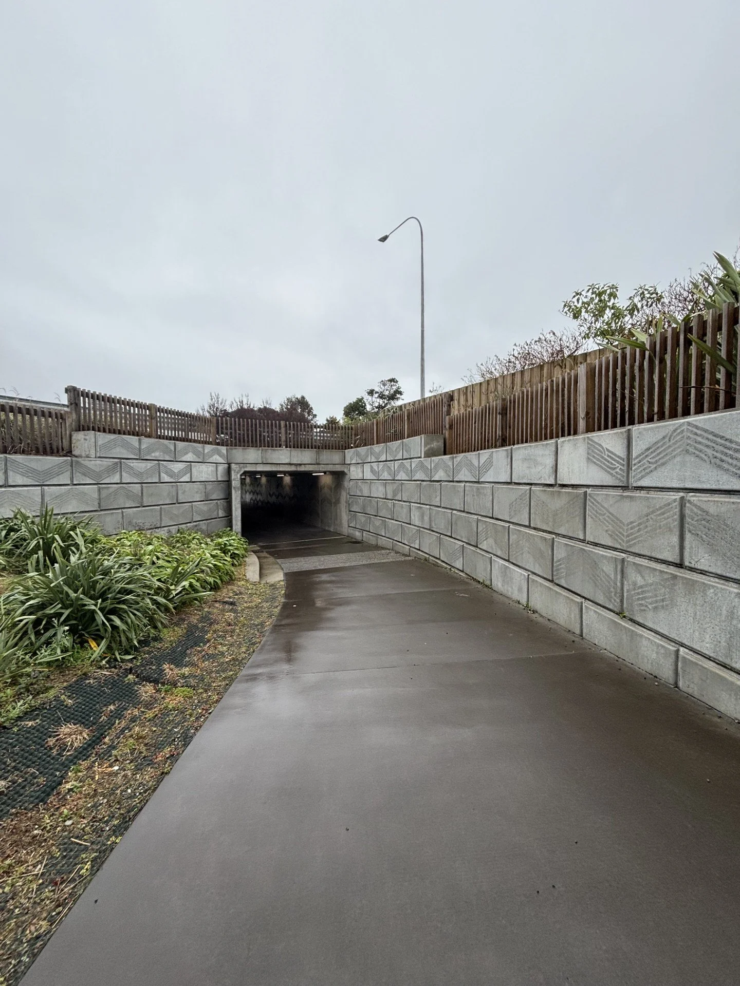 A concrete pathway leads to an underpass beneath a road, with a wooden fence on top of a stone retaining wall on the right side and greenery on the left, under an overcast sky.
