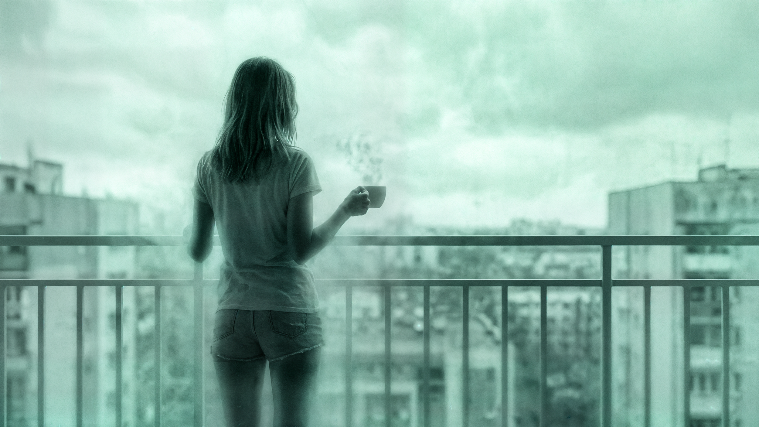 Woman with long hair in a T-shirt and shorts holding a coffee mug on a balcony, looking at cloudy sky and cityscape in the background.