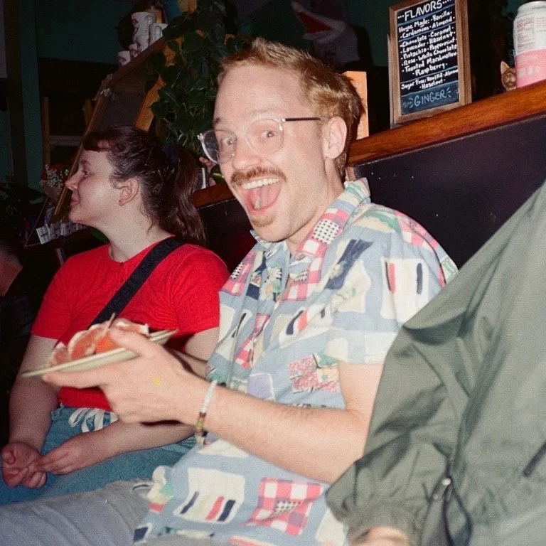 A man with glasses and a colorful, patterned short-sleeve shirt, smiling and holding a plate, sitting next to a woman in a red shirt, in a casual indoor setting.