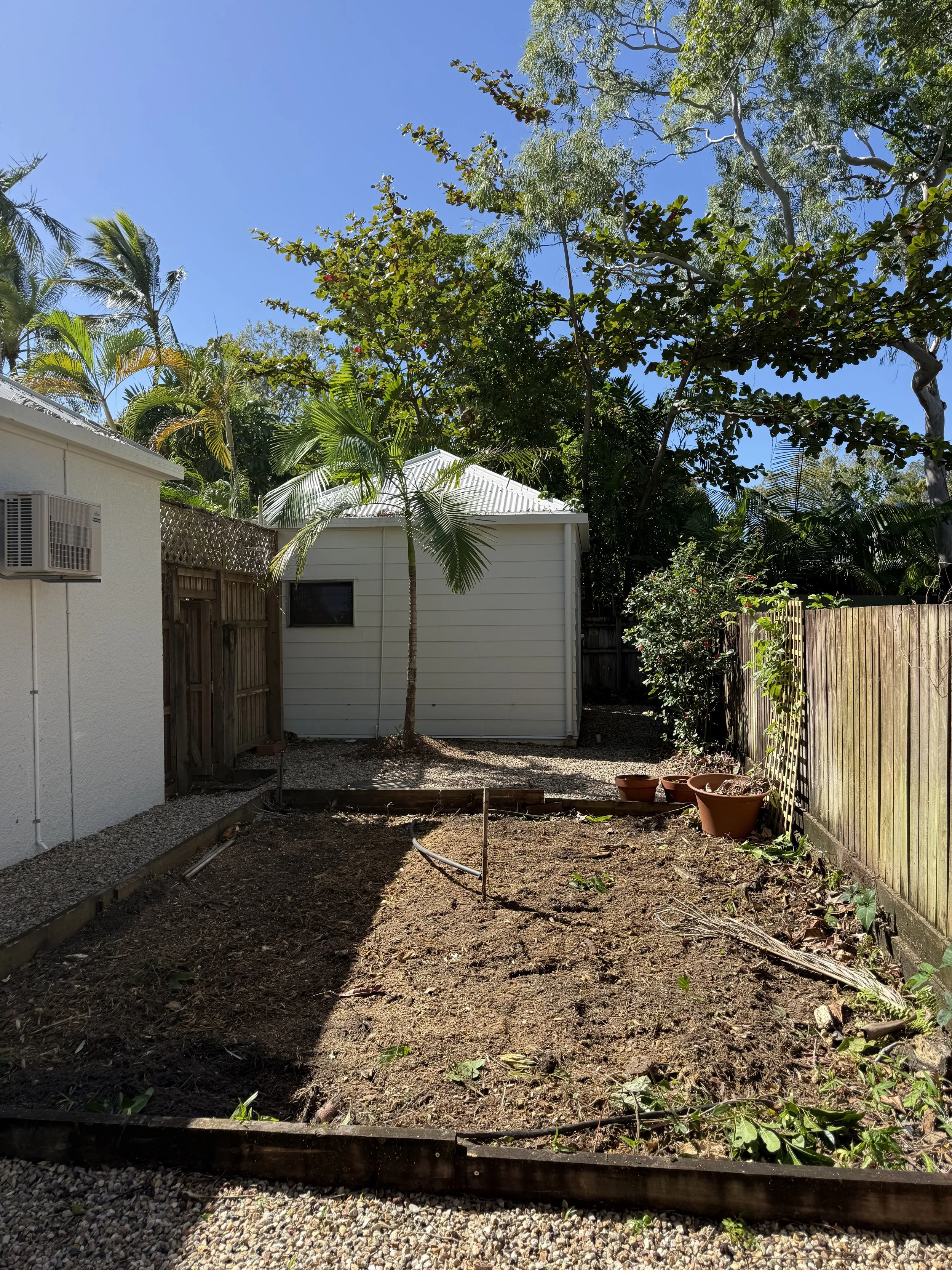 Backyard garden area with a recently tilled section of soil, empty plant pots, and surrounding trees and fencing.
