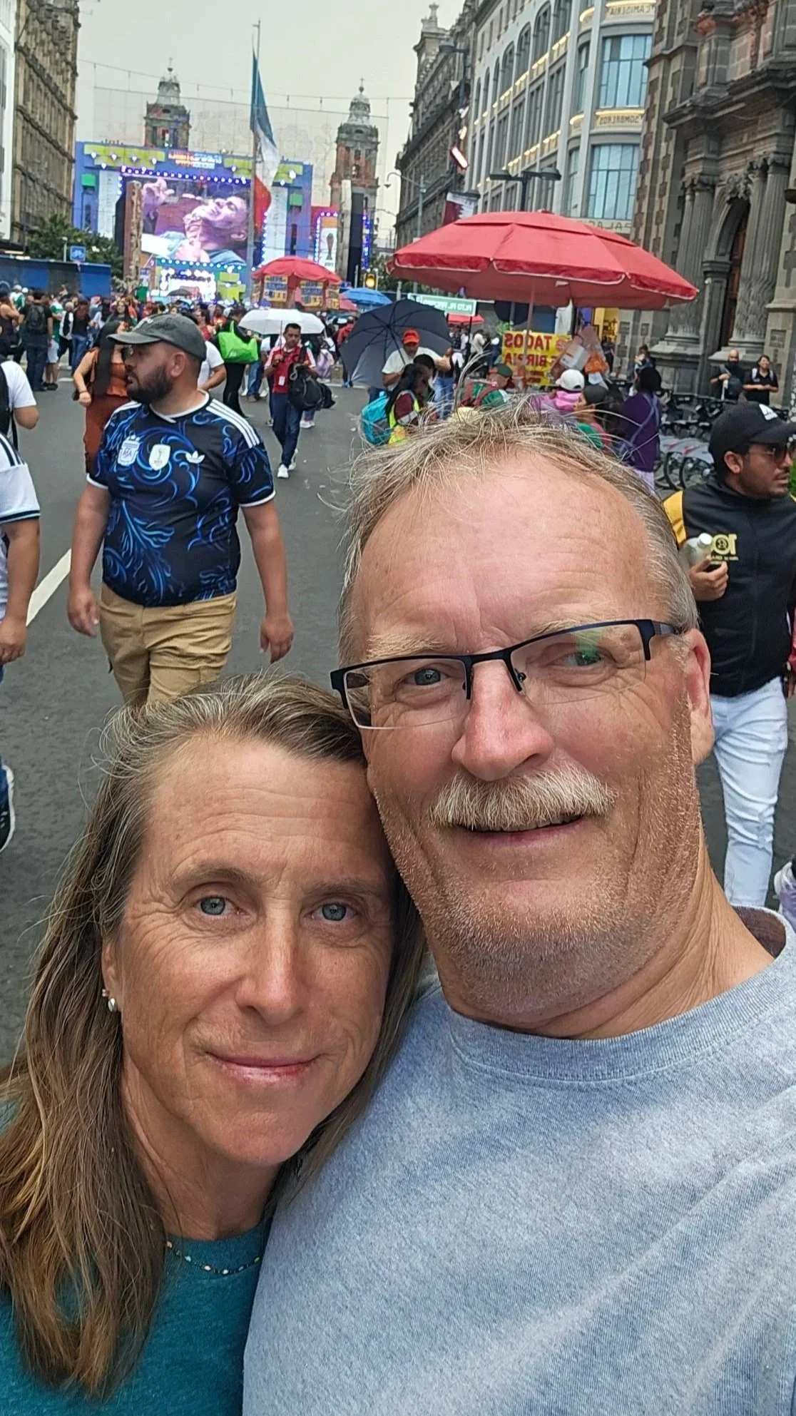 A selfie of a smiling older man with glasses and a woman with long hair in front of a crowd at an outdoor event in a city street.
