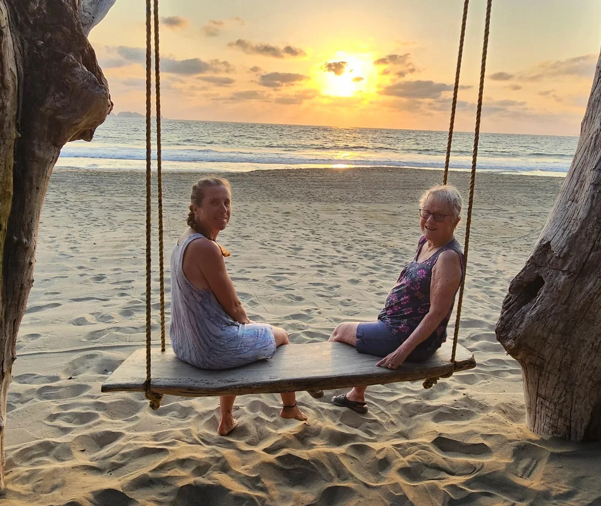 Two women sitting on a wooden swing at the beach during sunset, with ocean waves and a partly cloudy sky in the background.