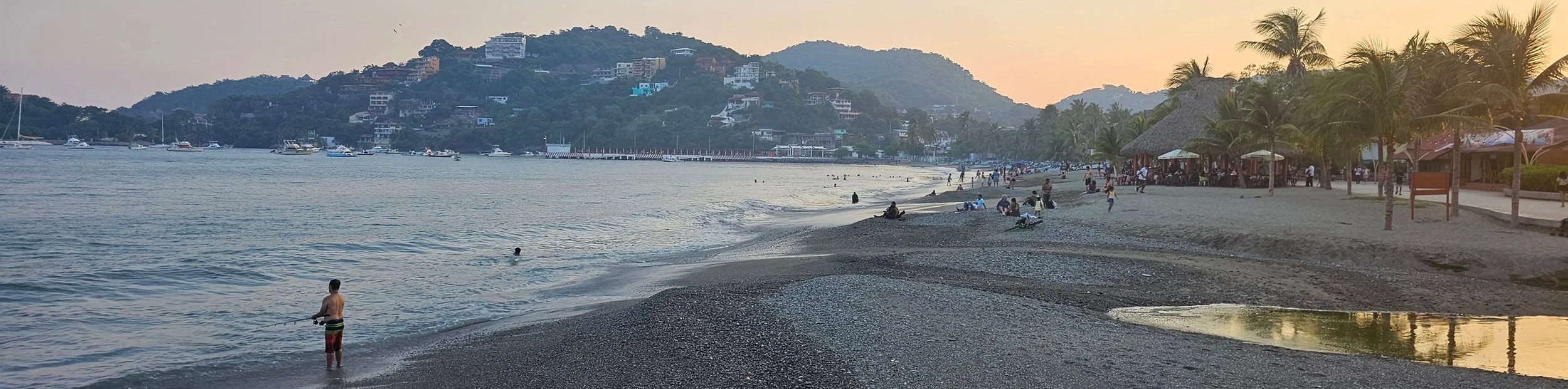 A beach scene at sunset with people relaxing on the sand and in the water, palm trees, and boats anchored offshore, with hillside houses in the background.