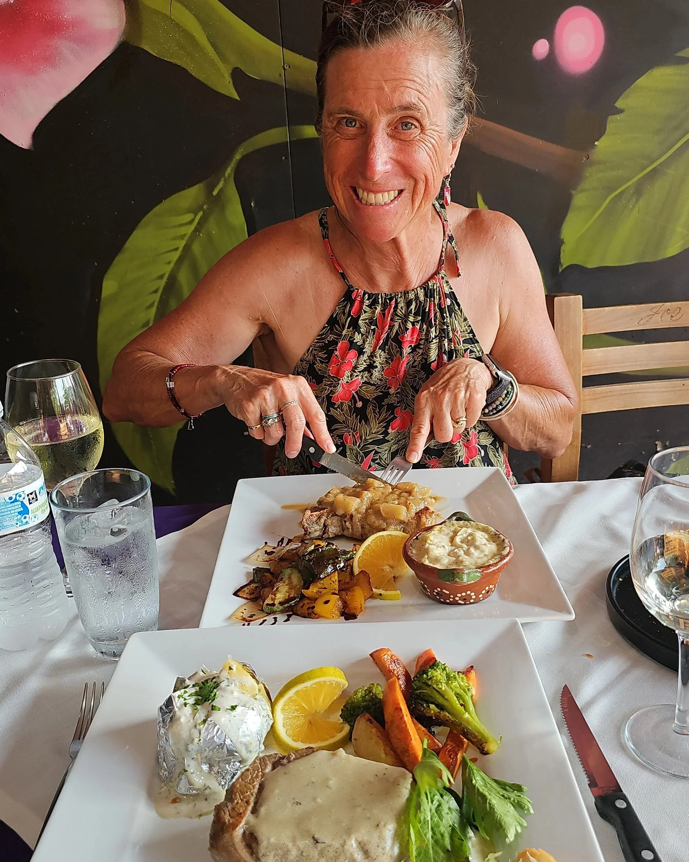 A smiling woman in a floral dress is cutting into a dish of cooked food at a dining table. The table has various dishes, including vegetables, a baked potato with sour cream, lemon slices, and a plate of grilled vegetables. There are glasses of water, silverware, and a black-handled knife on the table, with a decorative wall in the background.