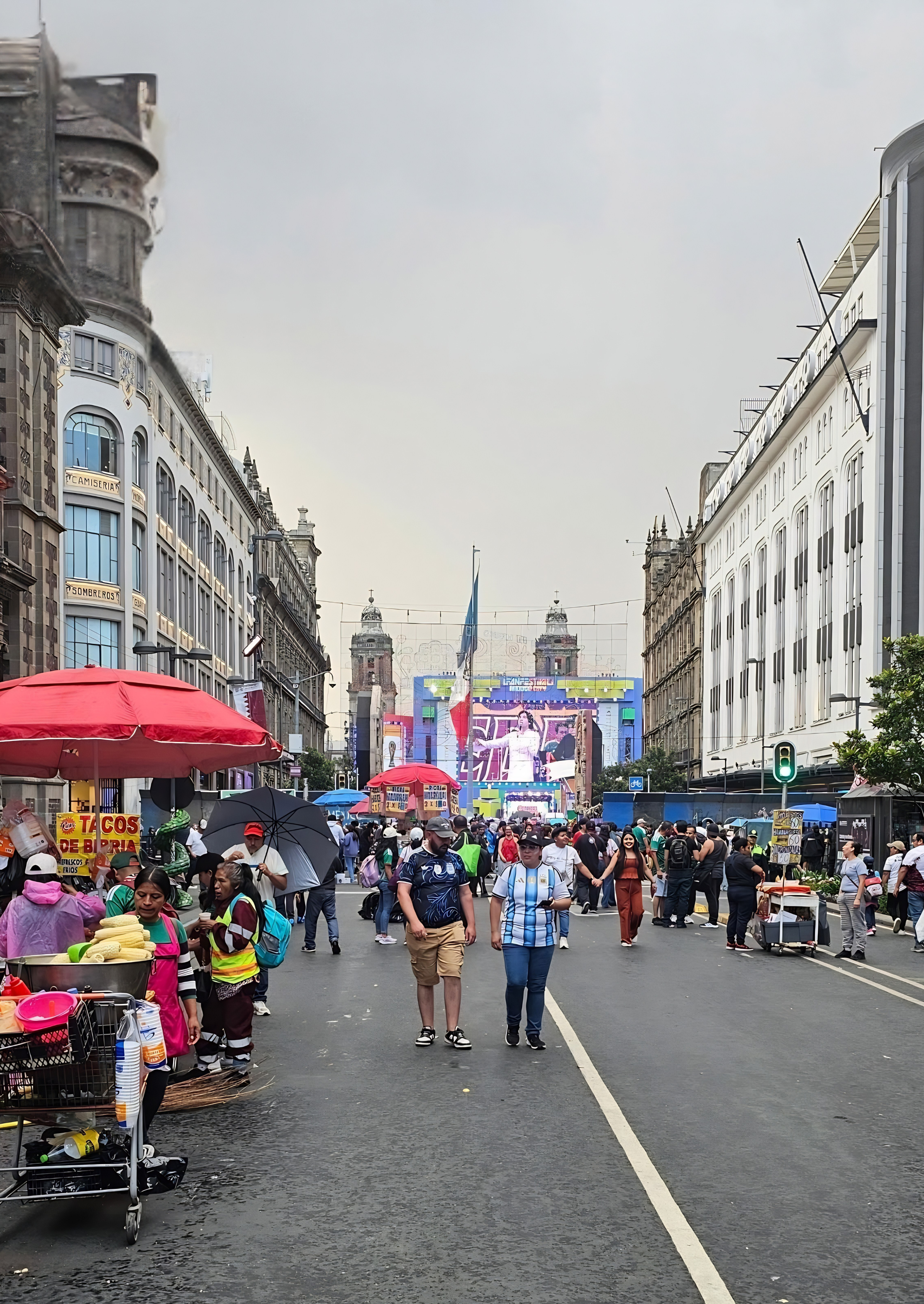 People walking on street with market stalls, umbrellas, and a large screen in the background showing a woman, with buildings on both sides and cloudy sky overhead.