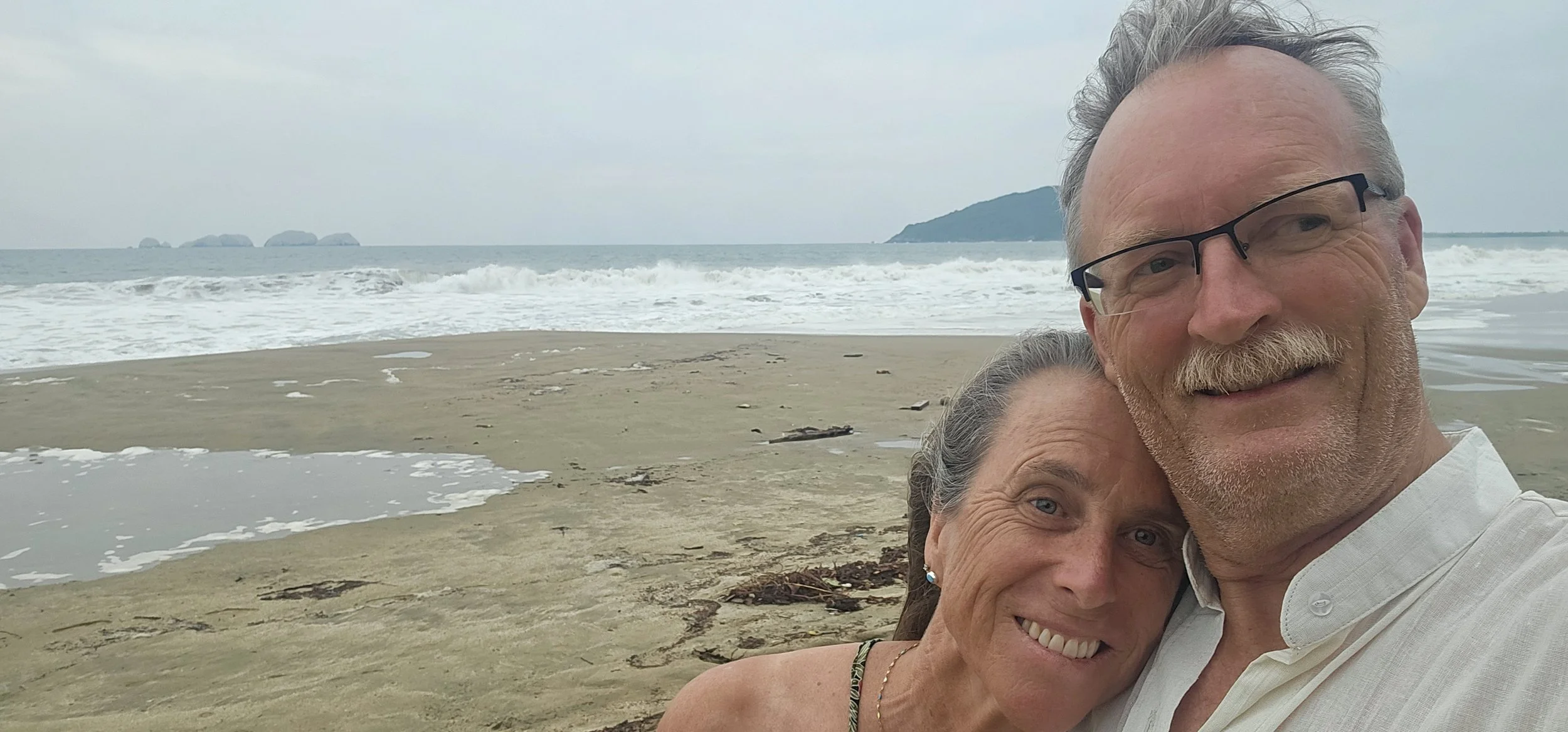 A smiling older couple taking a selfie on a beach with ocean waves, rocks, and a small island in the distance.