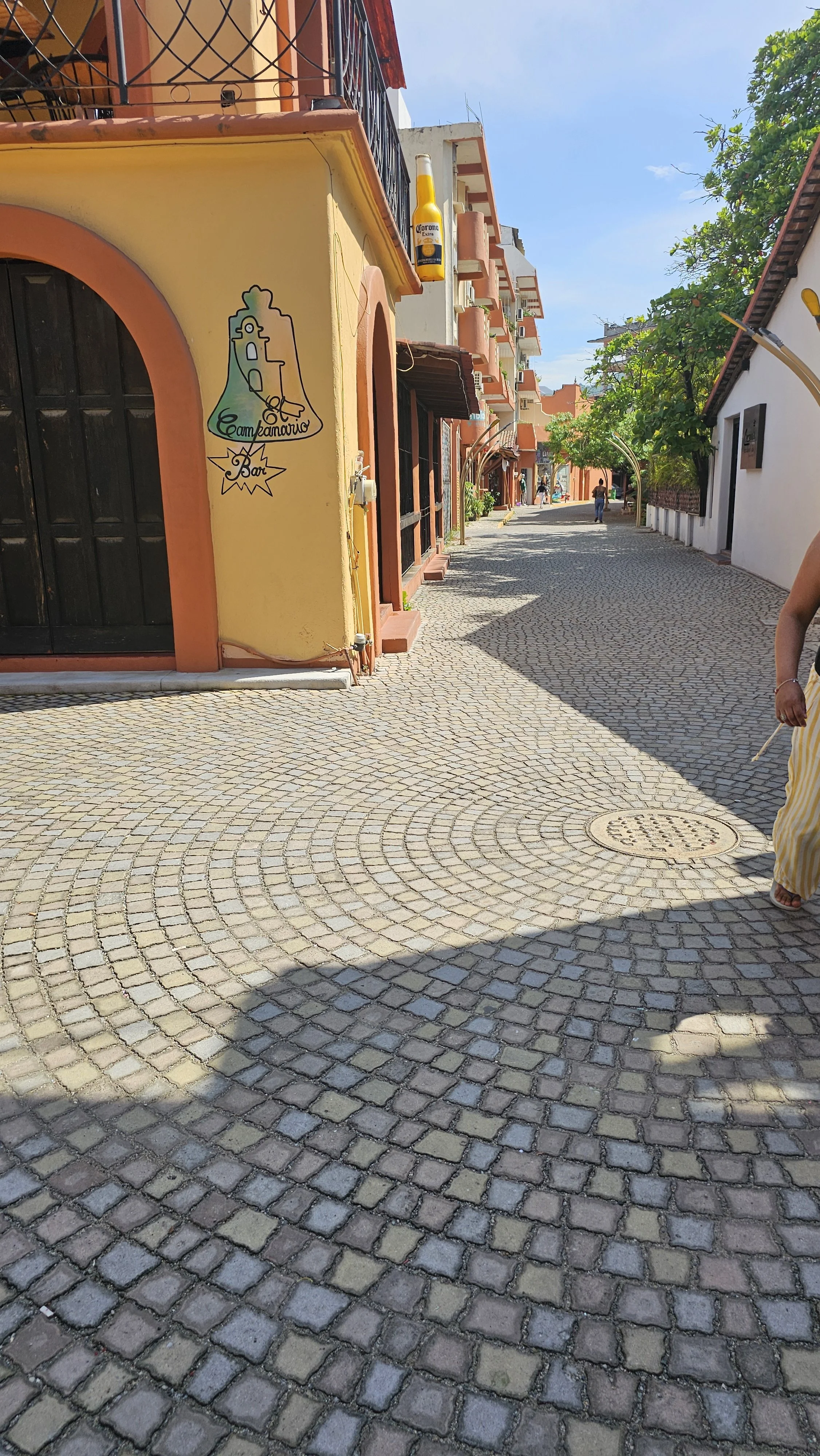 A cobblestone street in a colorful town with yellow, white, and orange buildings, some with balconies, trees, and a person in a striped yellow and white dress walking along the street.