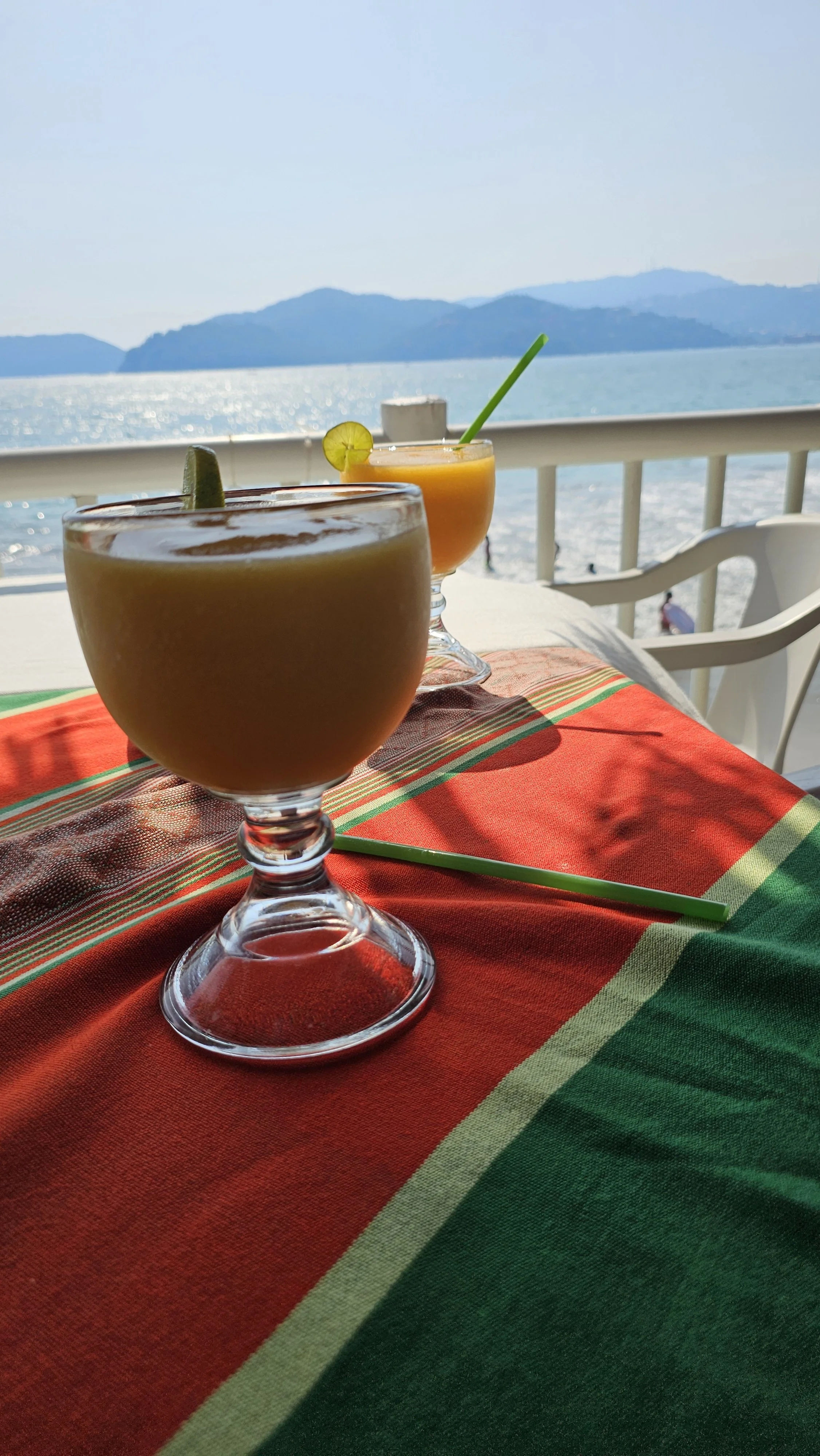 Two tropical drinks with lime slices and straws on a table with a red, green, and beige striped tablecloth, overlooking a body of water and distant mountains under a clear blue sky.