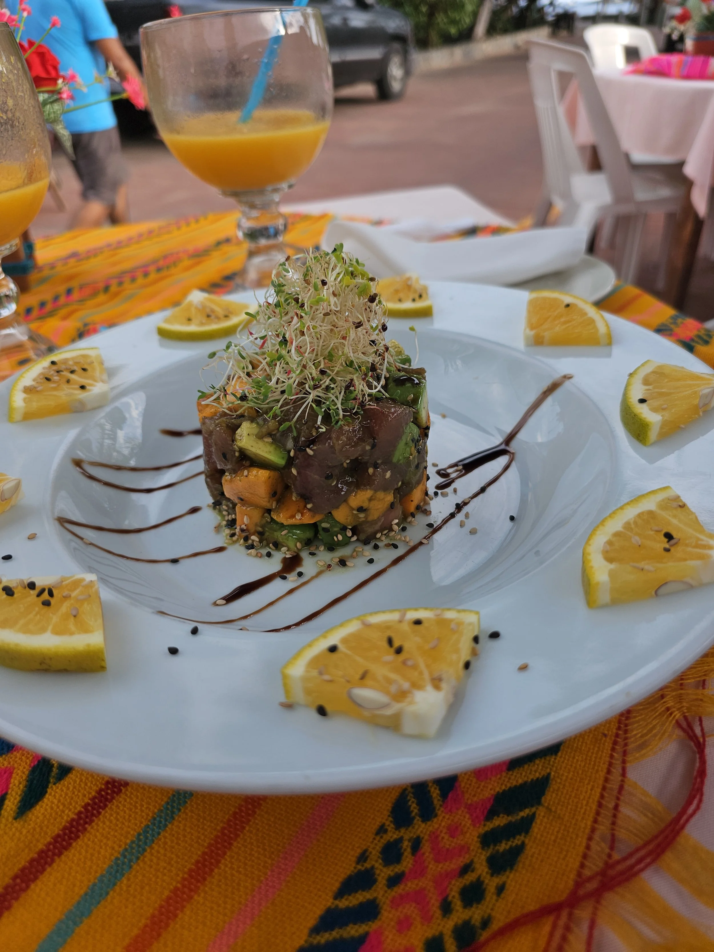 A plated dish with diced vegetables topped with sprouts, lemon wedges, and balsamic glaze, on a white plate on a colorful tablecloth. In the background, there are two glasses of orange juice and a white outdoor chair.