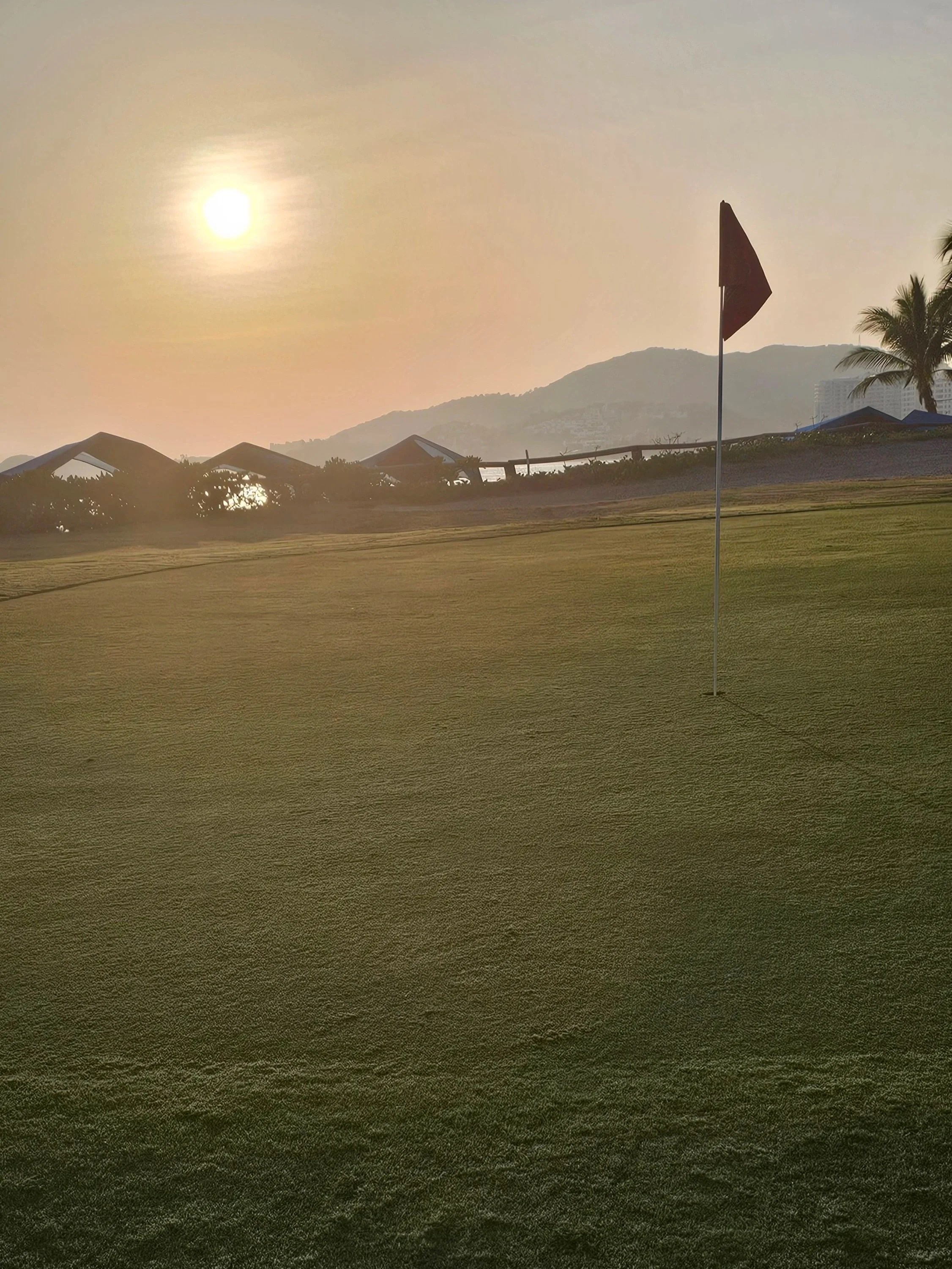 A golf course with a green, flag, nearby palm trees, tents or cabanas, a hillside, and a building in the distance, all under a bright sun in the sky.