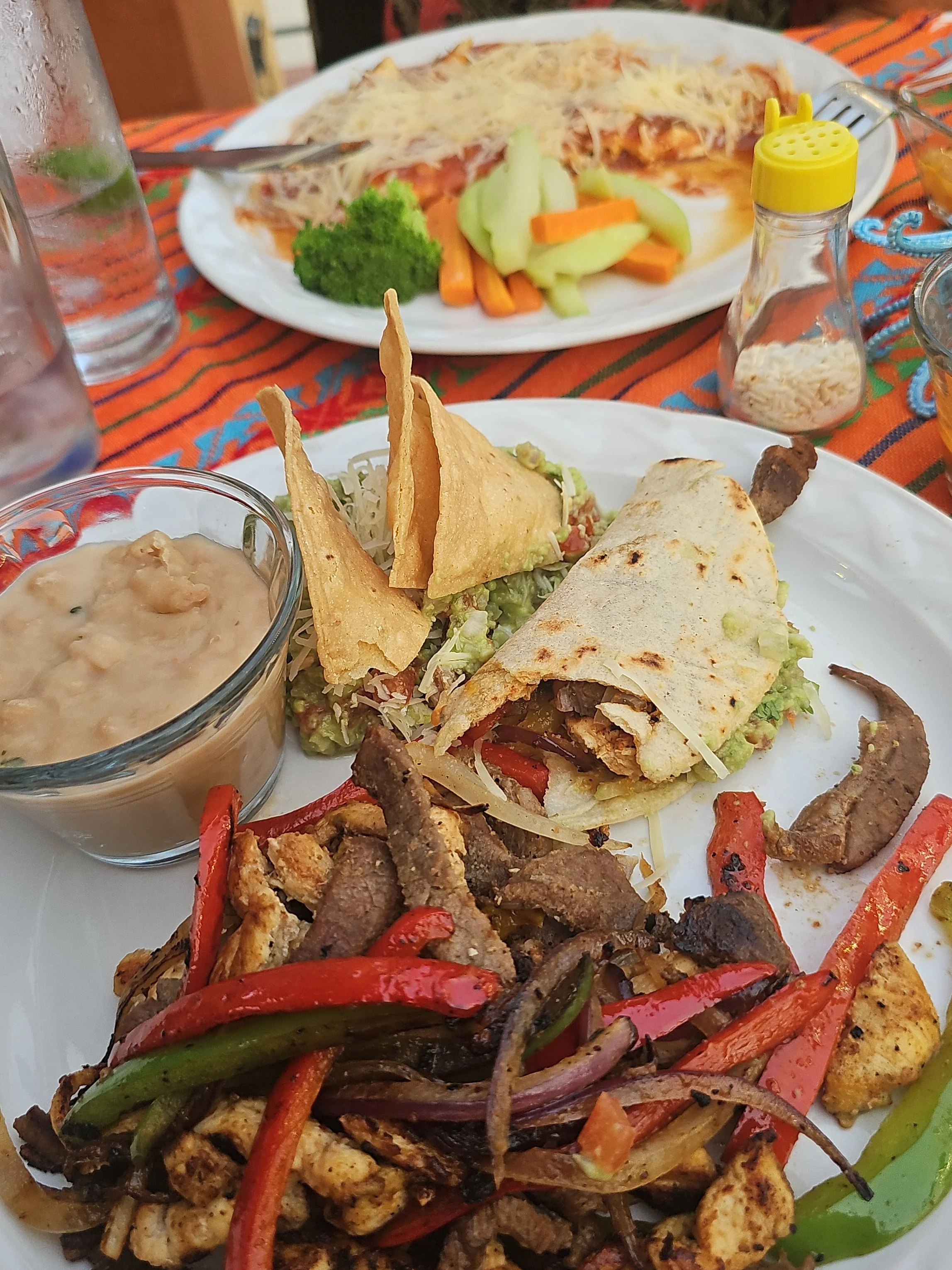 Plate of grilled meats and roasted vegetables, including red and green peppers, with a side of grilled chicken and seasoned beef, garnished with grilled vegetables and tortillas, served with a small bowl of sauce. In the background, another plate with pizza, cheese, and vegetables, and a container of grated cheese on a colorful tablecloth.