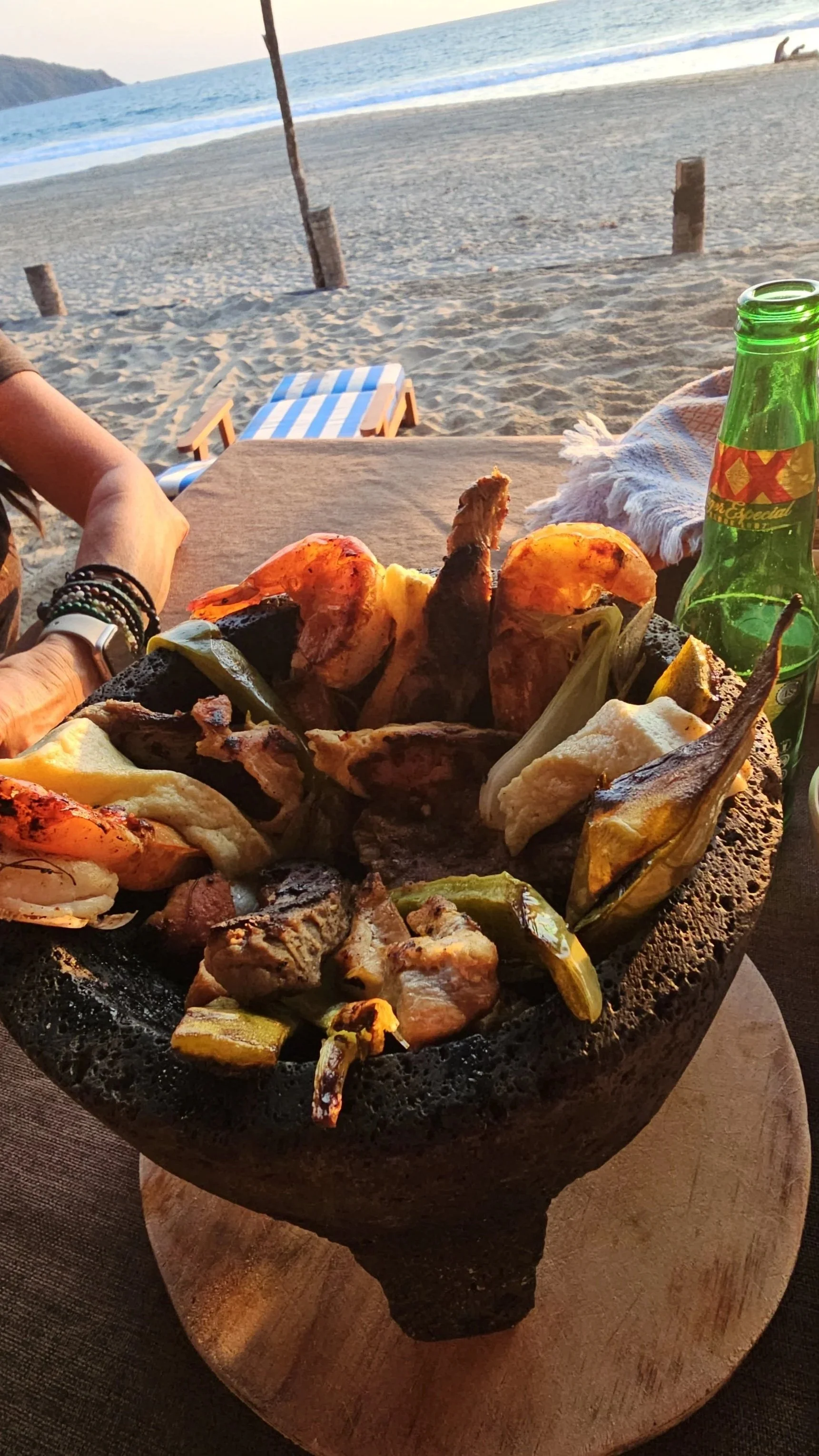 A table with grilled meats and vegetables in a volcanic stone bowl on a wooden serving board, with a beach and ocean in the background.
