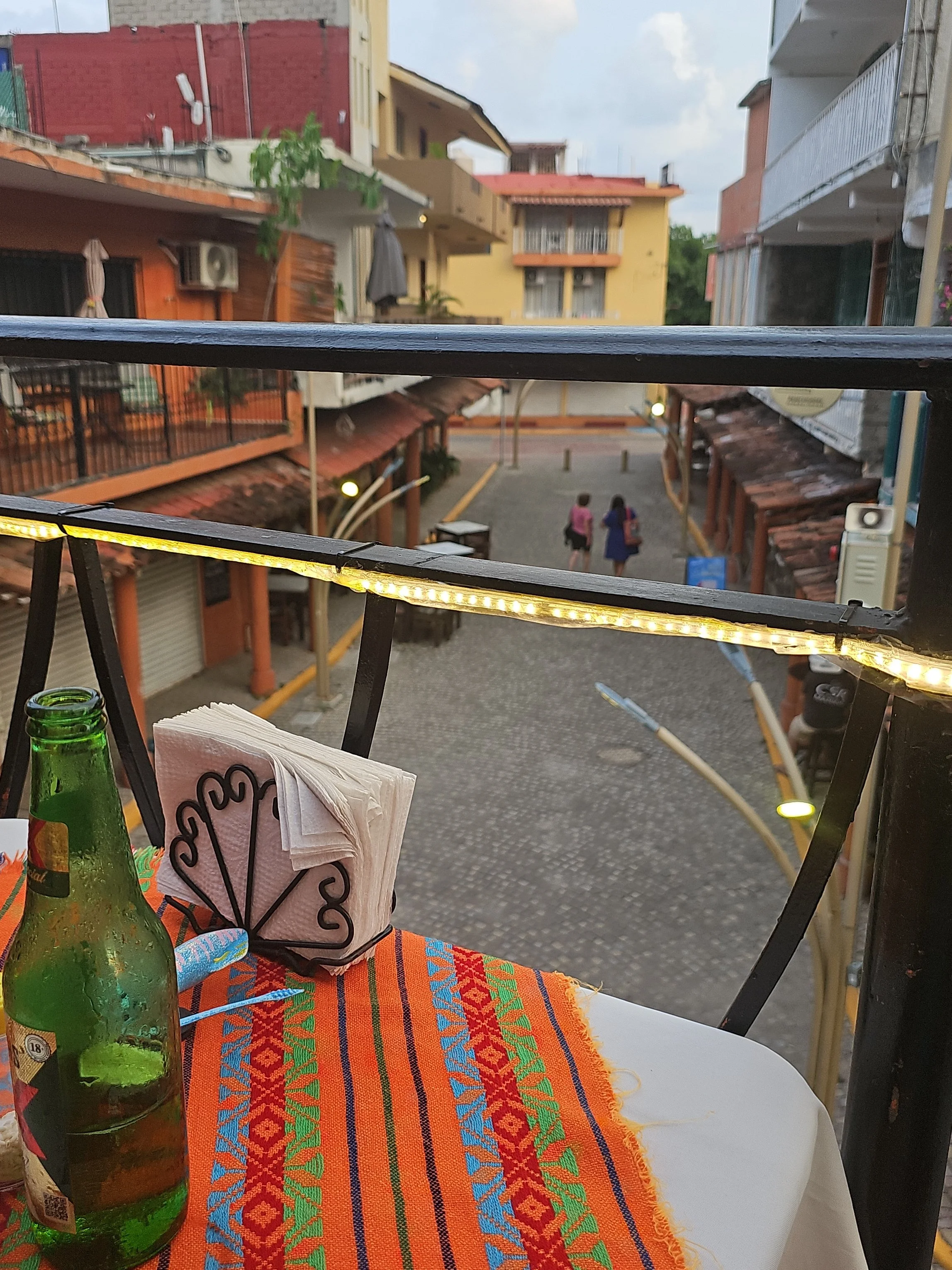 View of a small outdoor dining area at a restaurant or cafe, with a colorful patterned tablecloth, a green glass bottle, and paper napkins on the table. In the background, there are residential buildings with balconies, some with plants and balconies, and two people walking on the street.