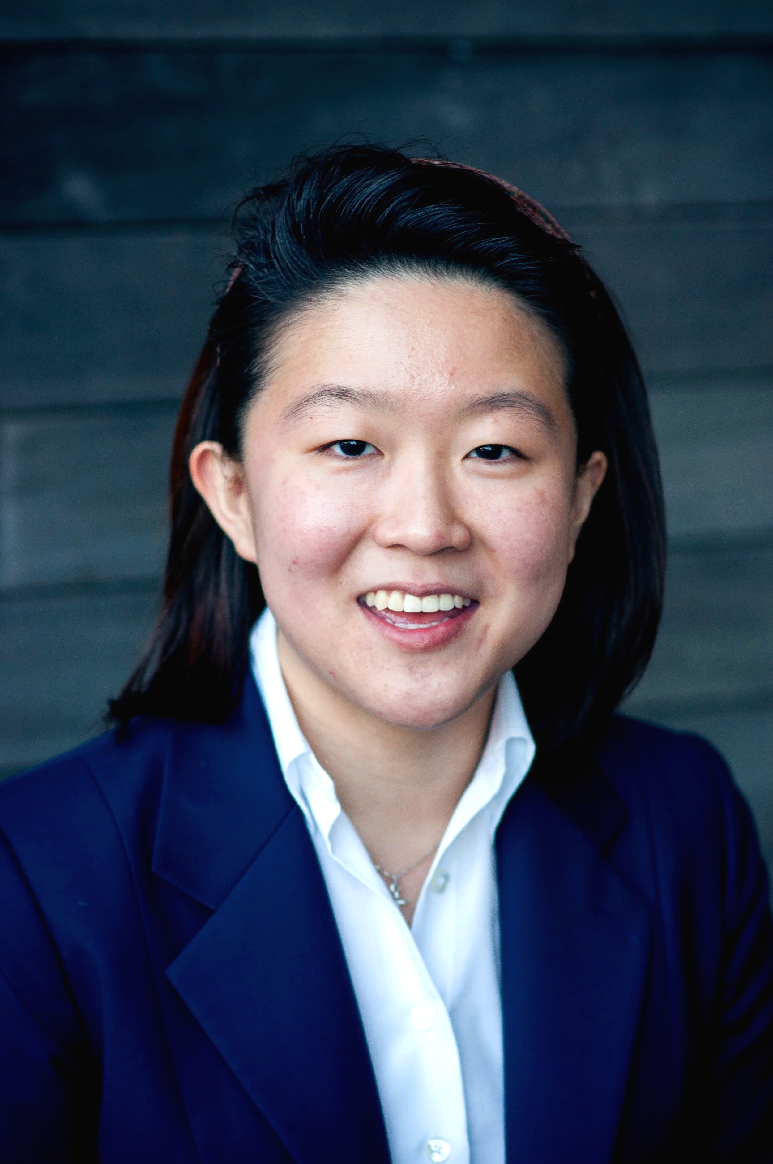 Close-up portrait of an Asian (Korean) woman with short black hair, smiling, wearing a navy blue blazer and a white shirt, against a dark background.