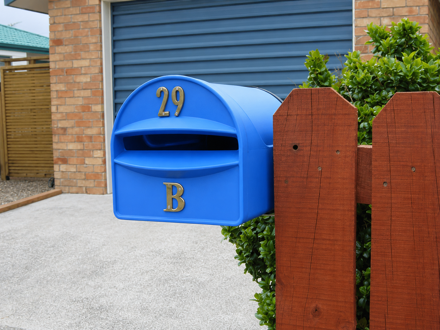 Blue mailbox with gold numbers '29' and letter 'B' on a wooden fence next to a brick house with a blue siding exterior.