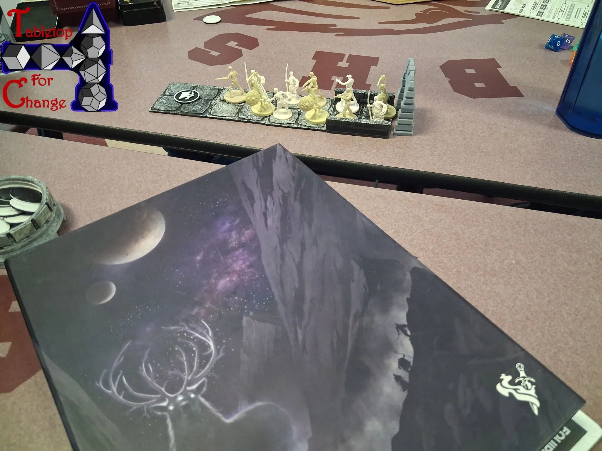 A tabletop with miniature fantasy figures arranged on a tiled passageway, surrounded by game pieces and a dice tray, during a tabletop role-playing game session.