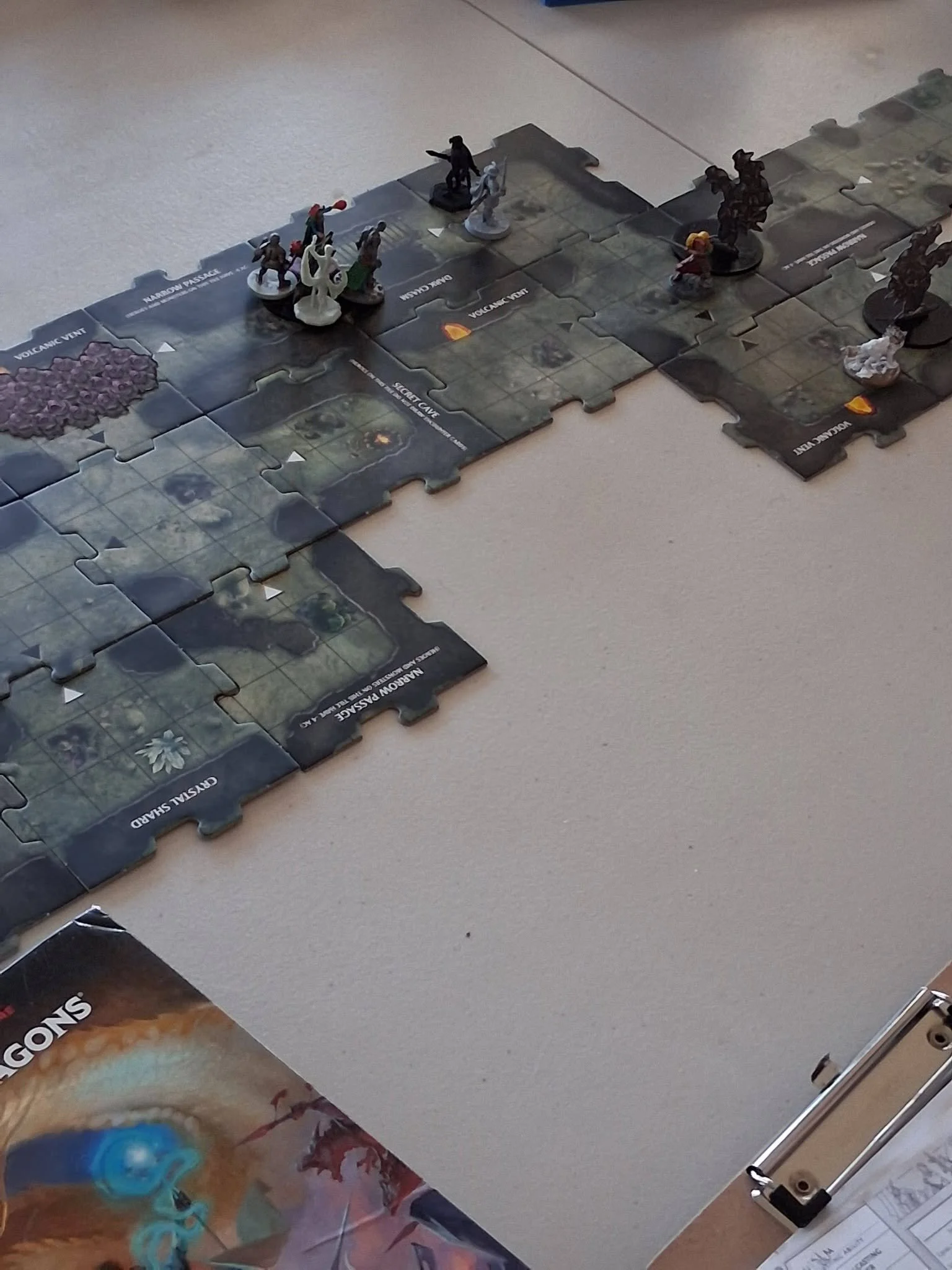 A tabletop game with puzzle tiles and miniature figurines representing characters and monsters, placed on a segmented game board with labeled areas, including 'Volcanic Vent' and 'Magma Passage'.