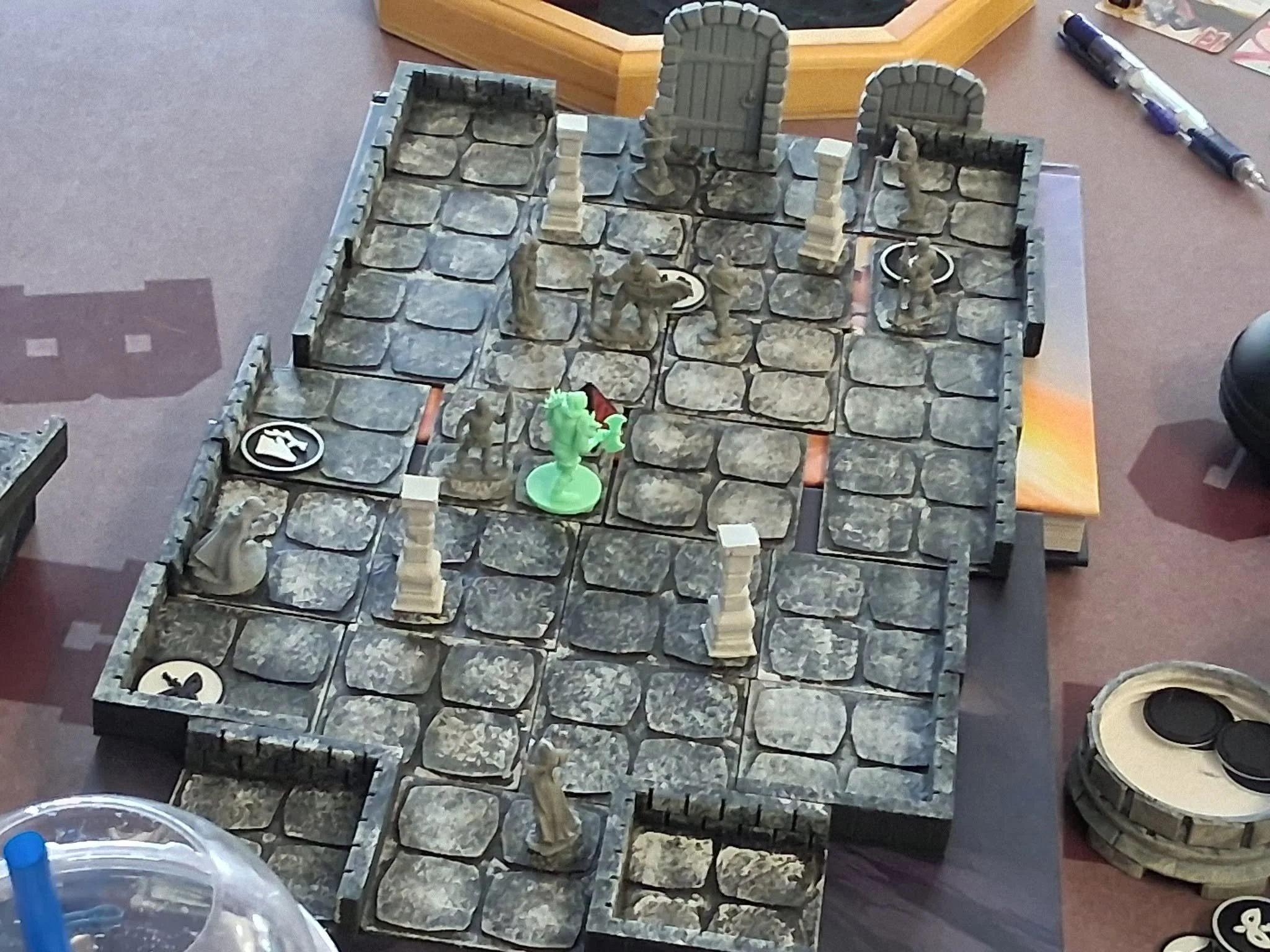 A tabletop game setup with a stone castle walls, two archways, and several miniature figurines of soldiers and a dragon. The game board shows pathways and character tokens, surrounded by gaming supplies and markers.