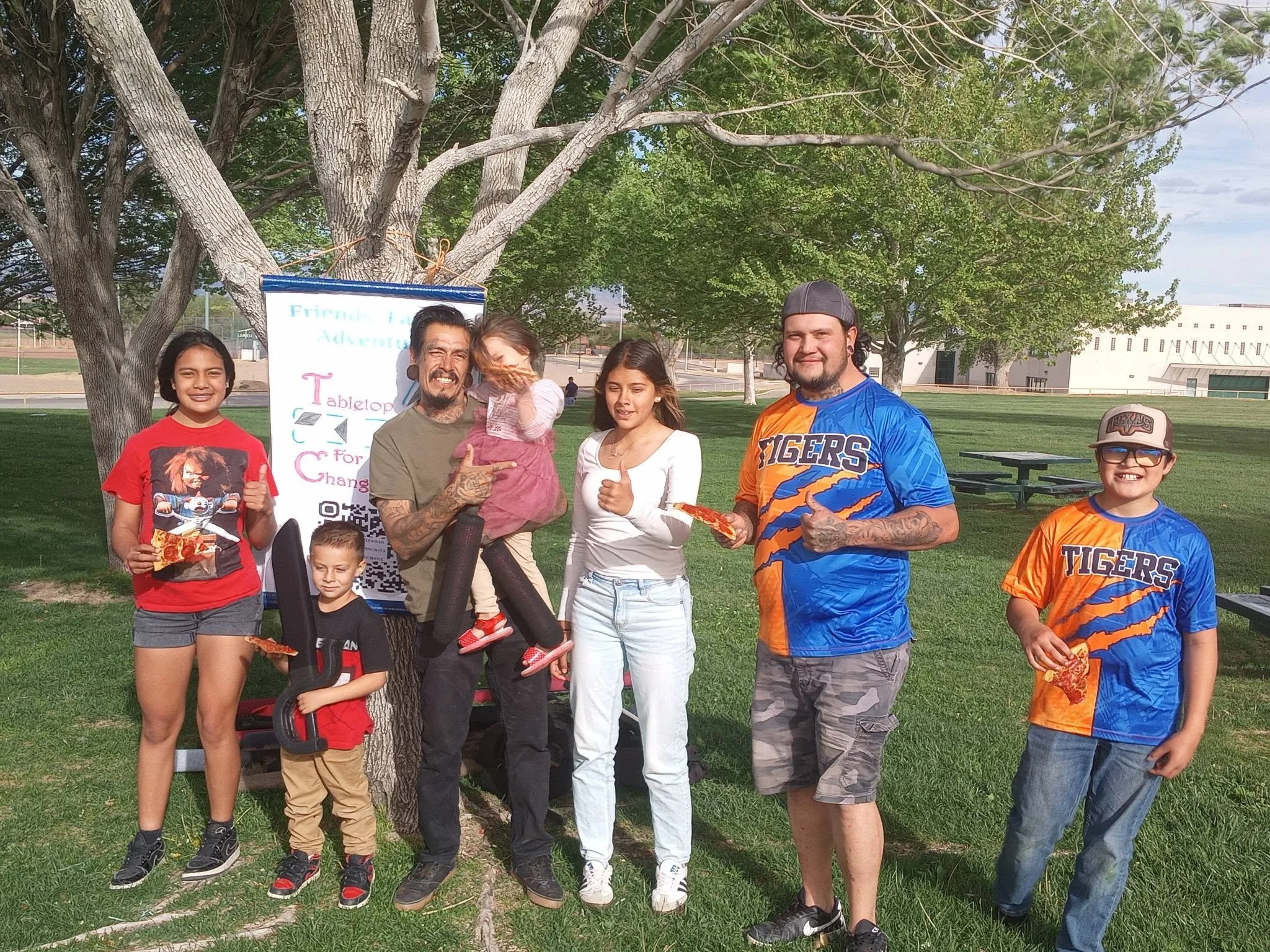 A group of seven diverse people, including children and adults, is gathered outdoors near a tree in a park on a sunny day. They are smiling, giving thumbs up, and holding pizza slices.
