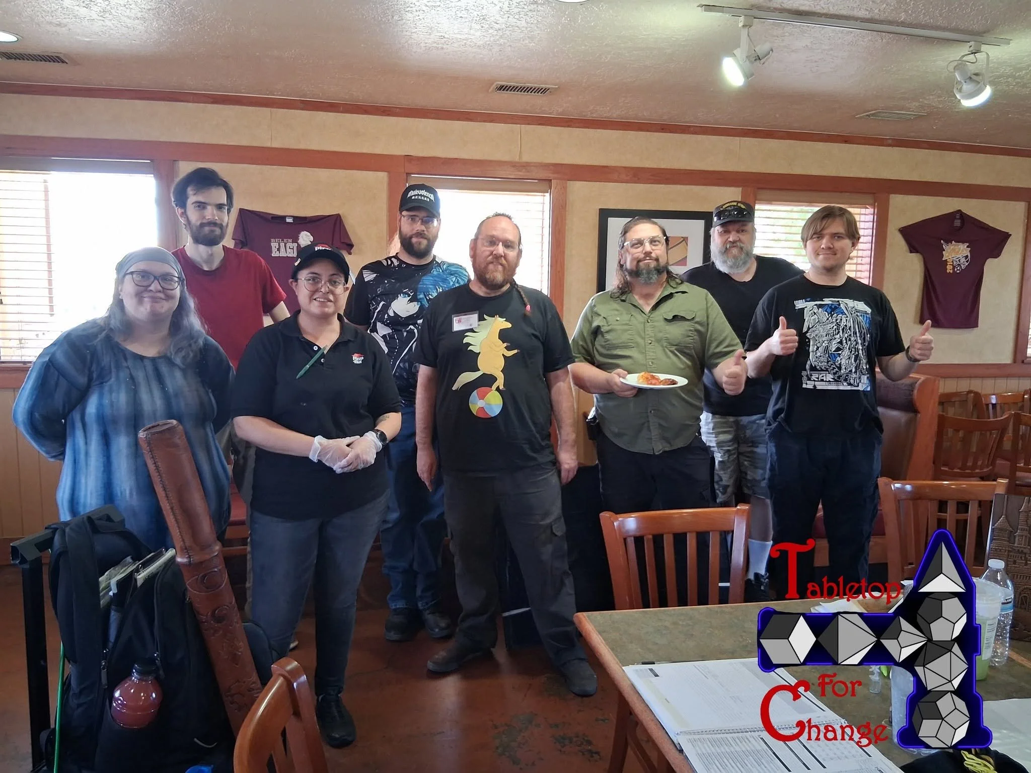 Group of nine people standing inside a restaurant, with two maroon t-shirts hanging on the wall behind them. One person is holding a plate with food, and another is giving a thumbs-up gesture. A man in the middle has a black t-shirt with a pegasus design. The group appears to be posing for a photo, with some smiling and others looking serious.