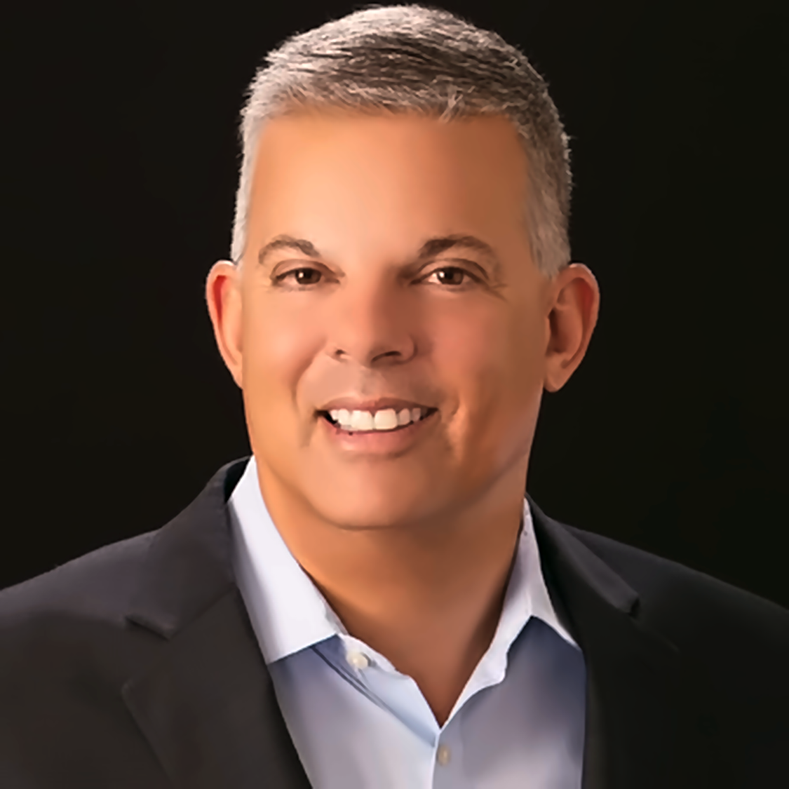Professional headshot of a smiling man with short gray hair, wearing a black suit jacket and a light blue collared shirt against a black background.