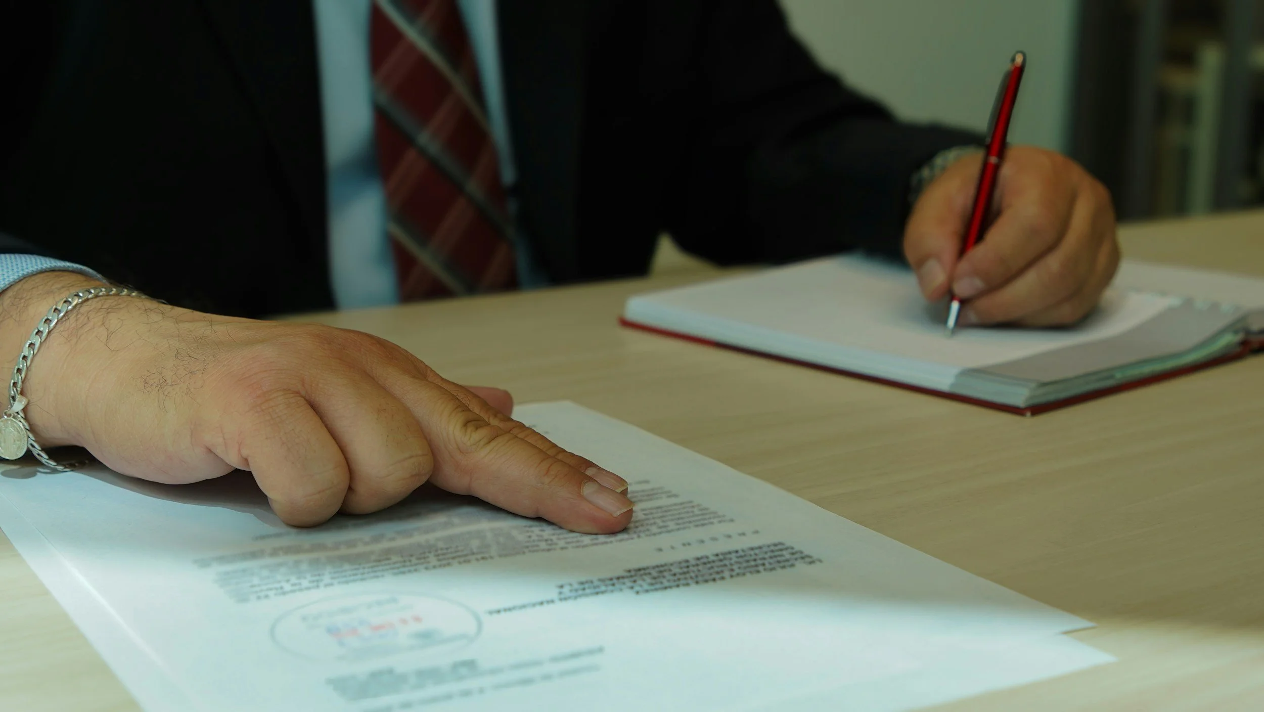 A person is sitting at a desk, pointing at a document with their finger, while holding a red pencil in their other hand. The person is wearing a black jacket, a blue shirt, a red and blue striped tie, a silver bracelet, and a silver watch. There is an open book on the desk.