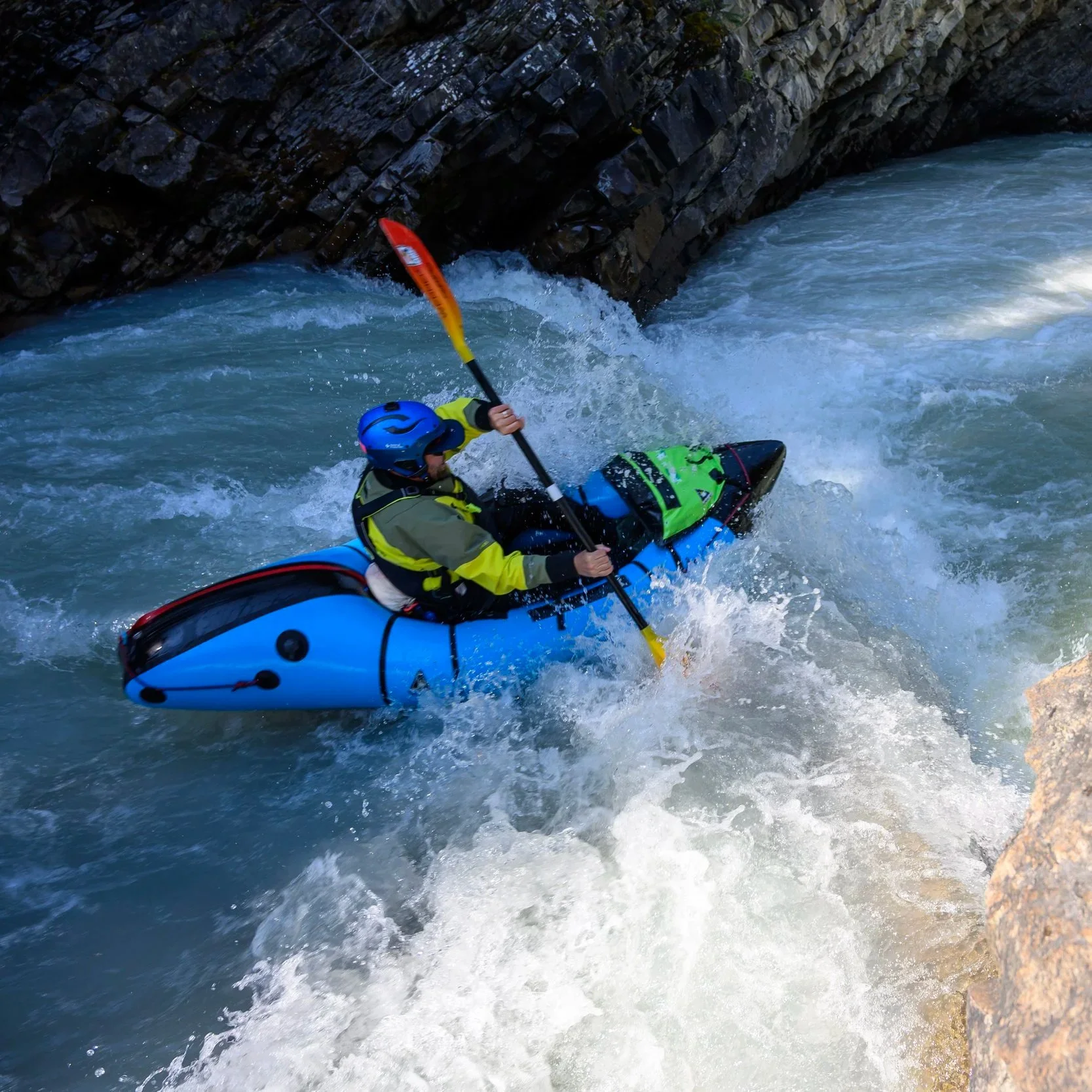A person wearing a blue helmet and yellow life jacket kayaking in white water rapids near a rocky cliff side.