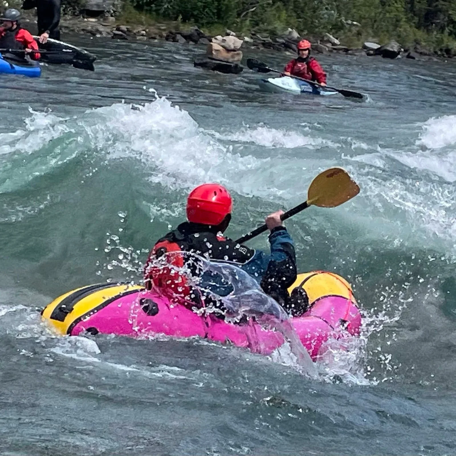 People white water kayaking with helmets and life jackets, navigating a river with rocks and splashing water.