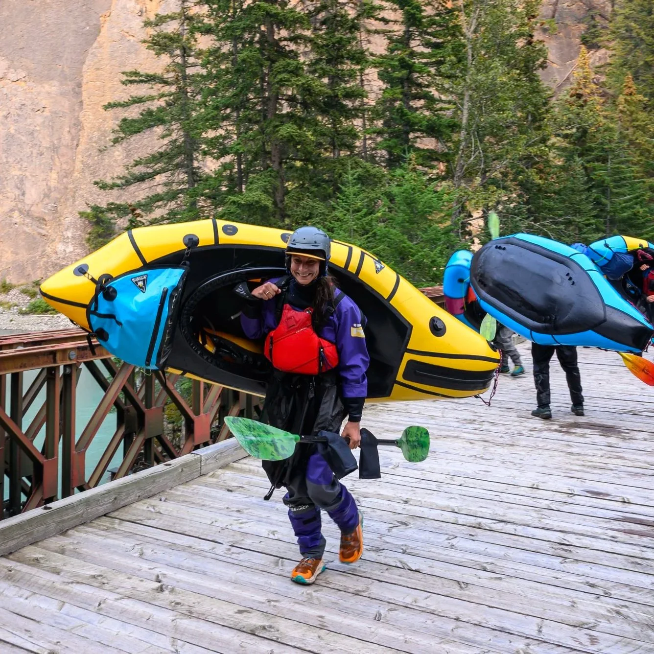 Woman in kayaking gear walking on a wooden bridge, carrying a yellow kayak with blue bags attached, in a forested area.