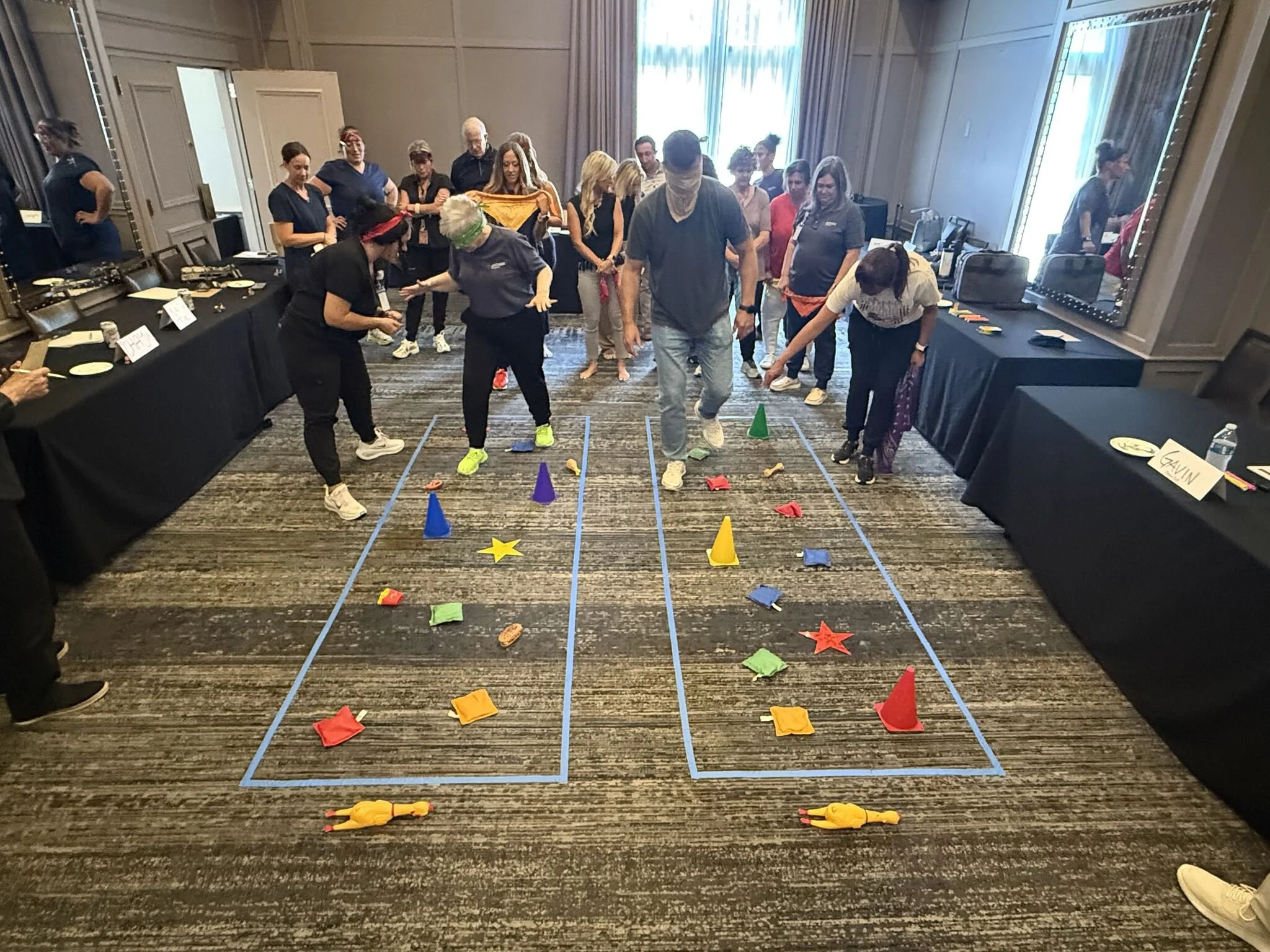 A group of people participating in an indoor team-building activity involving stepping on colored fabric pieces in a marked grid on the floor, with some spectators watching.