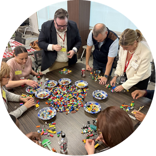 People gathered around a table playing with colorful Lego bricks, some stacking and sorting them, with bowls of additional Lego pieces in the center.