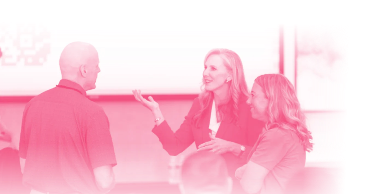Three people in a conversation, a man and two women, in a professional setting, with a presentation screen in the background.