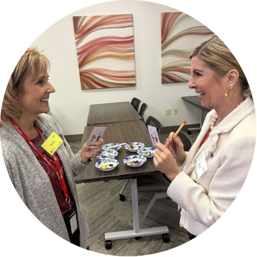 Two women smiling and chatting in an office setting, exchanging small cards, with decorated plates on a table behind them.