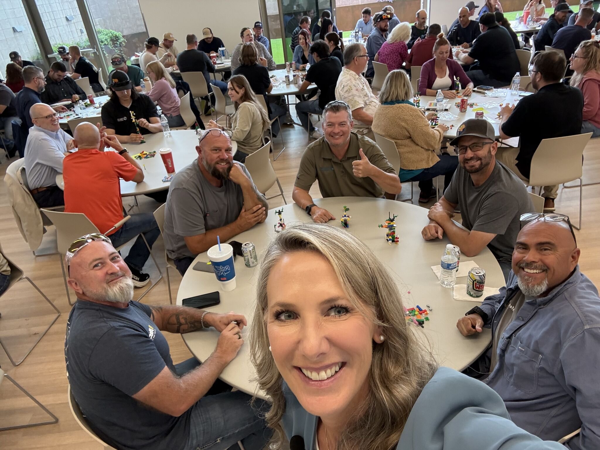 Heather Hodges taking a selfie at a LEGO® Serious Play® team workshop, with attendees gathered at tables engaging with the activity.