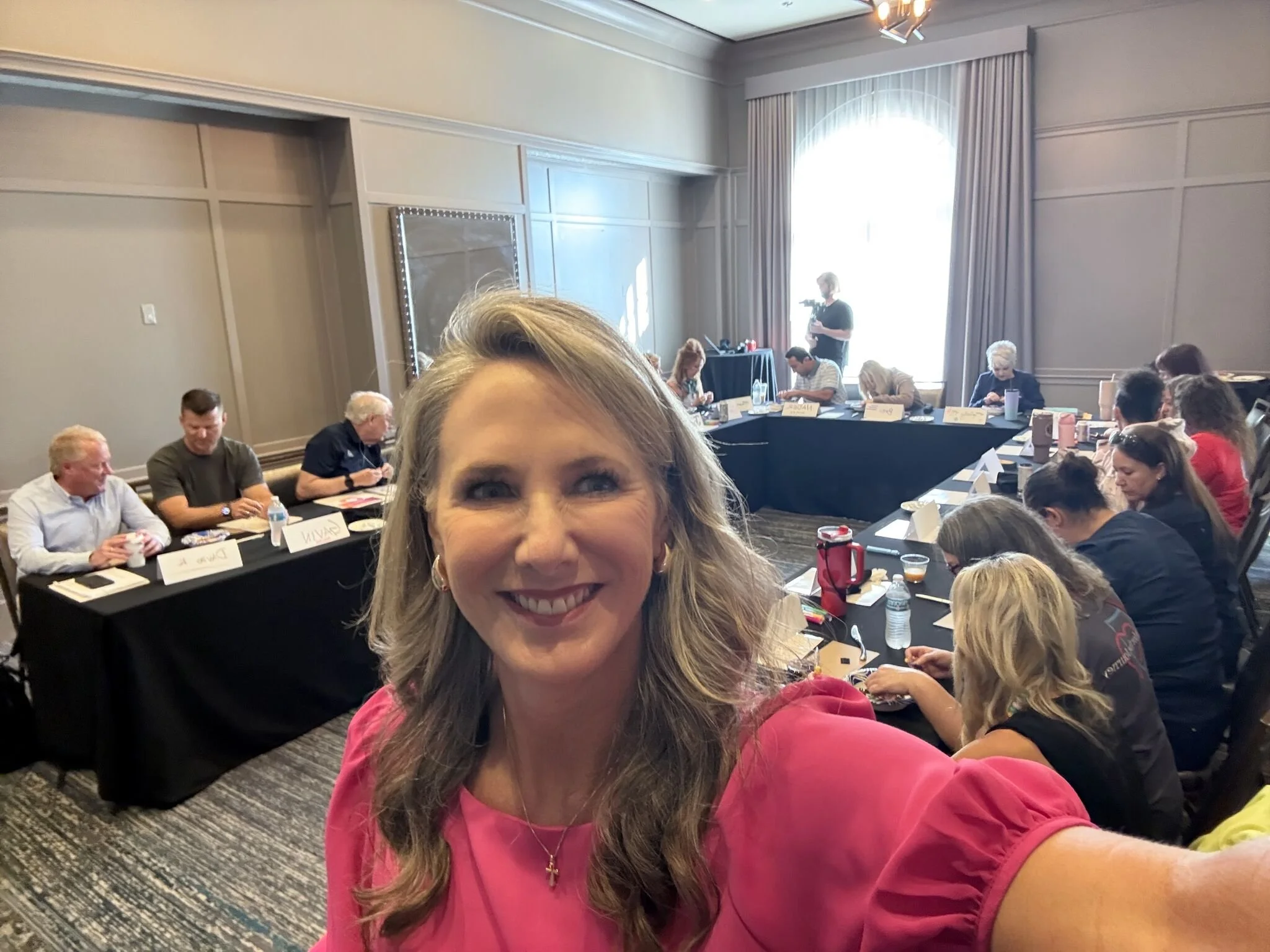 Heather Hodges taking a selfie at a conference, with attendees seated at tables in the background.