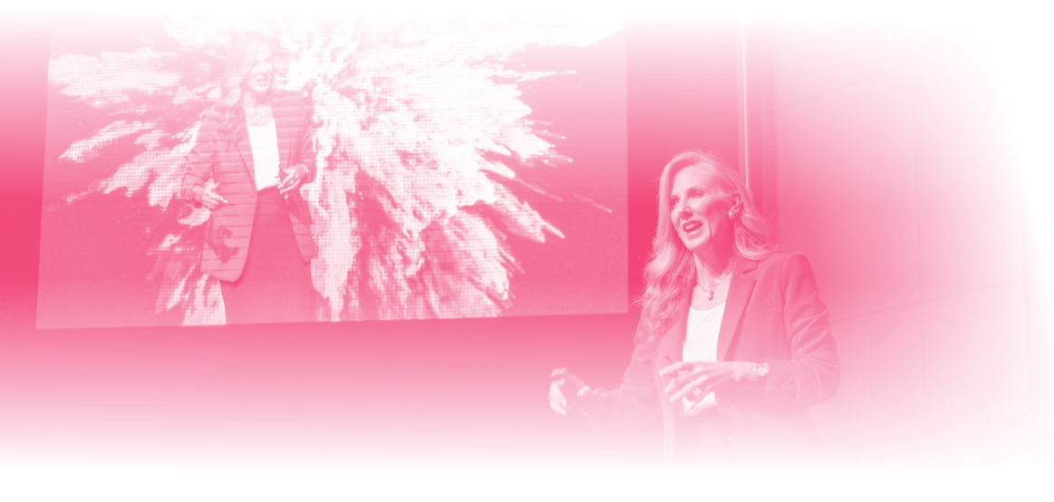 A woman is giving a presentation on stage with a large screen behind her displaying her image.